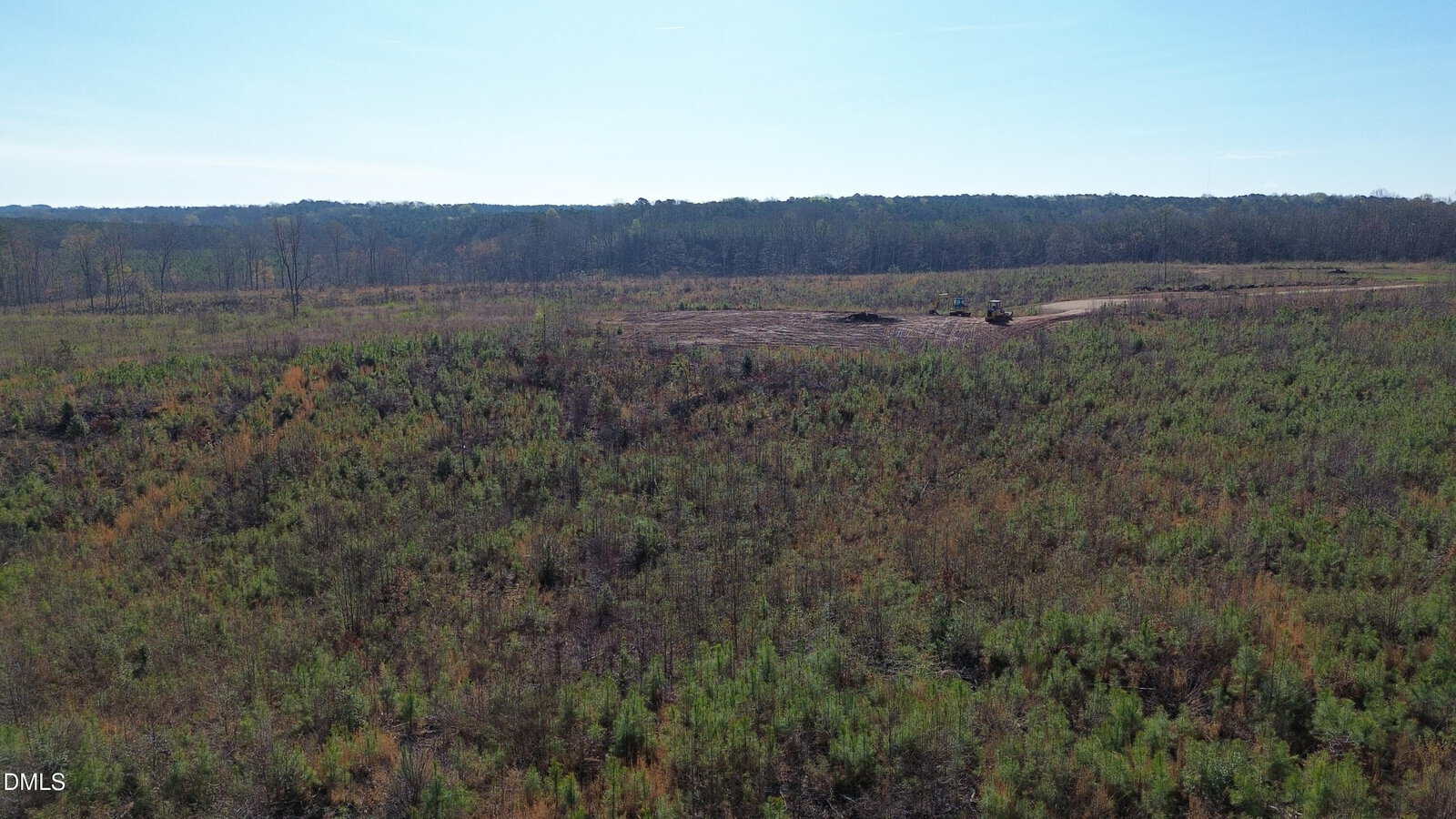 Aerial view of a large, open field with sparse vegetation and distant trees under a clear blue sky; a small piece of machinery is visible near the center.