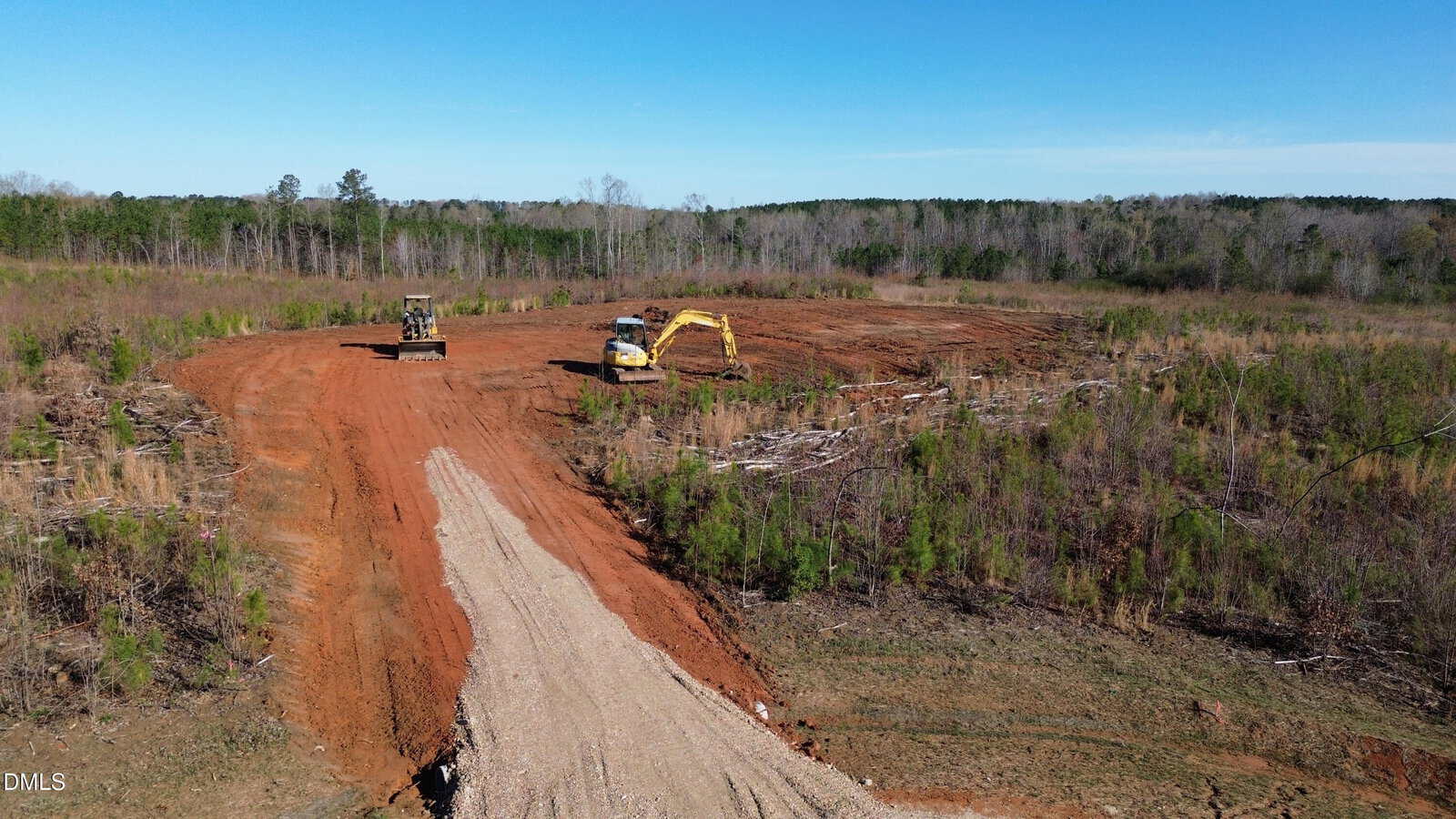 A construction site with cleared red dirt, two excavators working, and sparse vegetation surrounding the area. A dirt road leads into the site, bordered by small green bushes and trees.