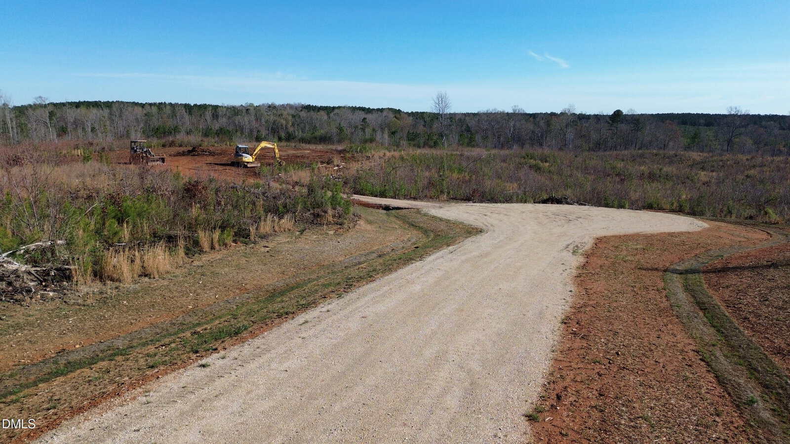 A dirt road curves through a mostly barren landscape with sparse vegetation near 175 Tillery Drive in Louisburg. In the distance, a yellow excavator is working on a patch of cleared land under a clear blue sky.