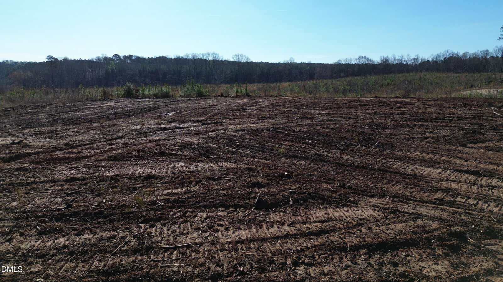 A large, cleared area of land with tire tracks and sparse vegetation; a tree line is visible in the background under a clear blue sky.