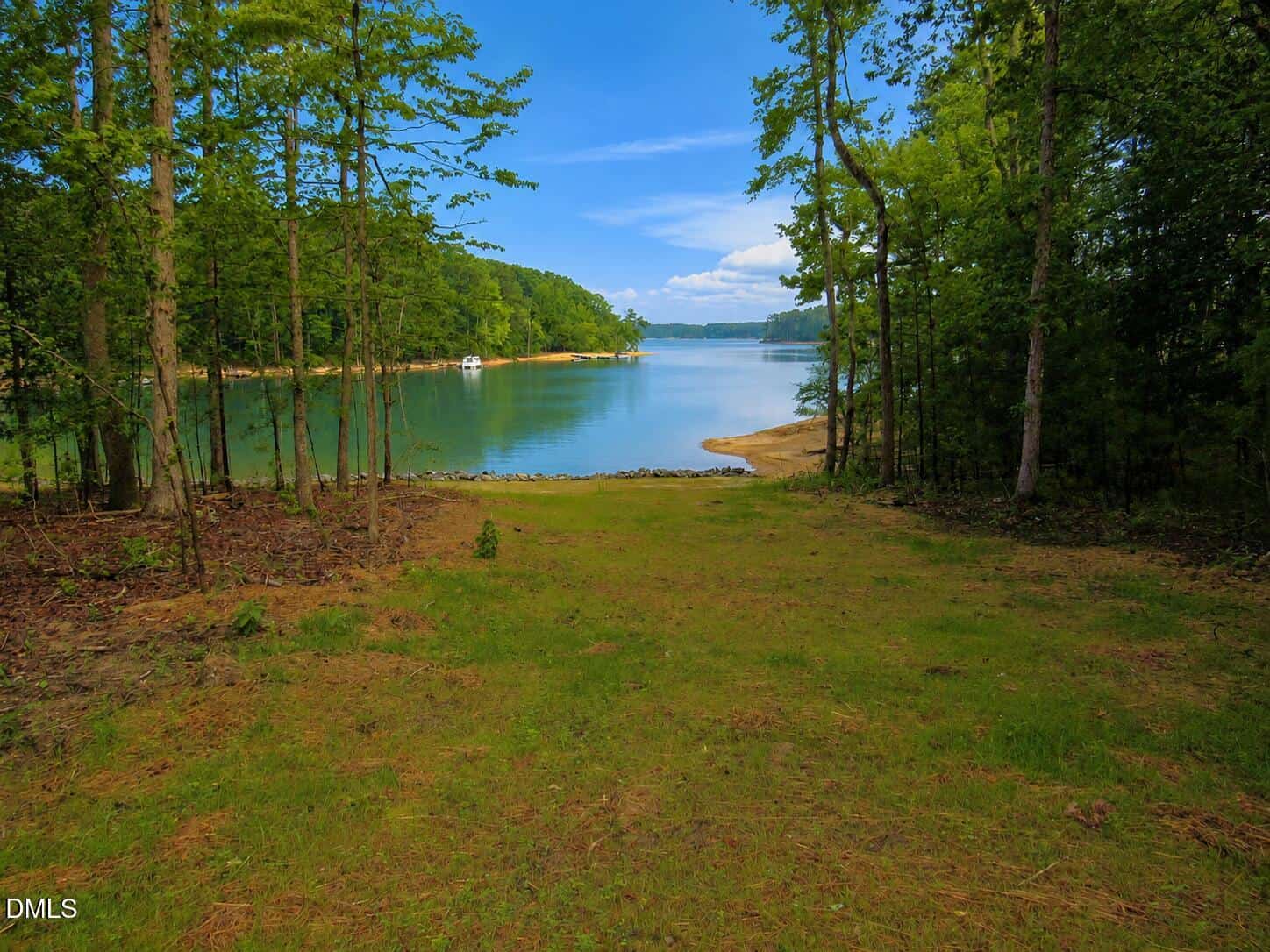 A grassy clearing leads to a calm lake surrounded by trees, with a sandy shoreline and blue sky with scattered clouds in the background.