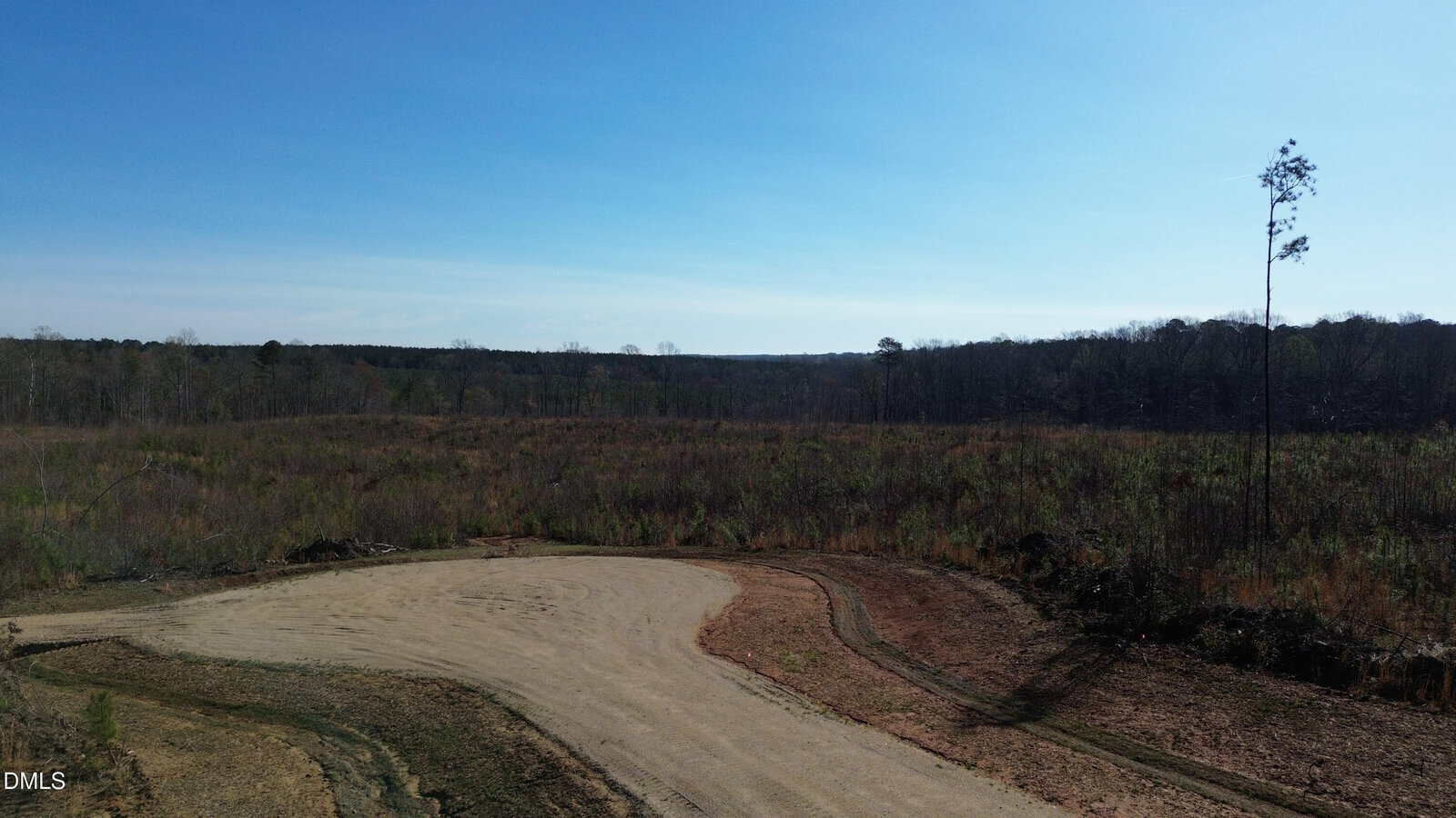 A dirt road curves through a mostly barren and grassy field under a clear blue sky, bordered by sparse trees and forest in the background, capturing the tranquil rural charm near 160 Tillery Drive in Louisburg.