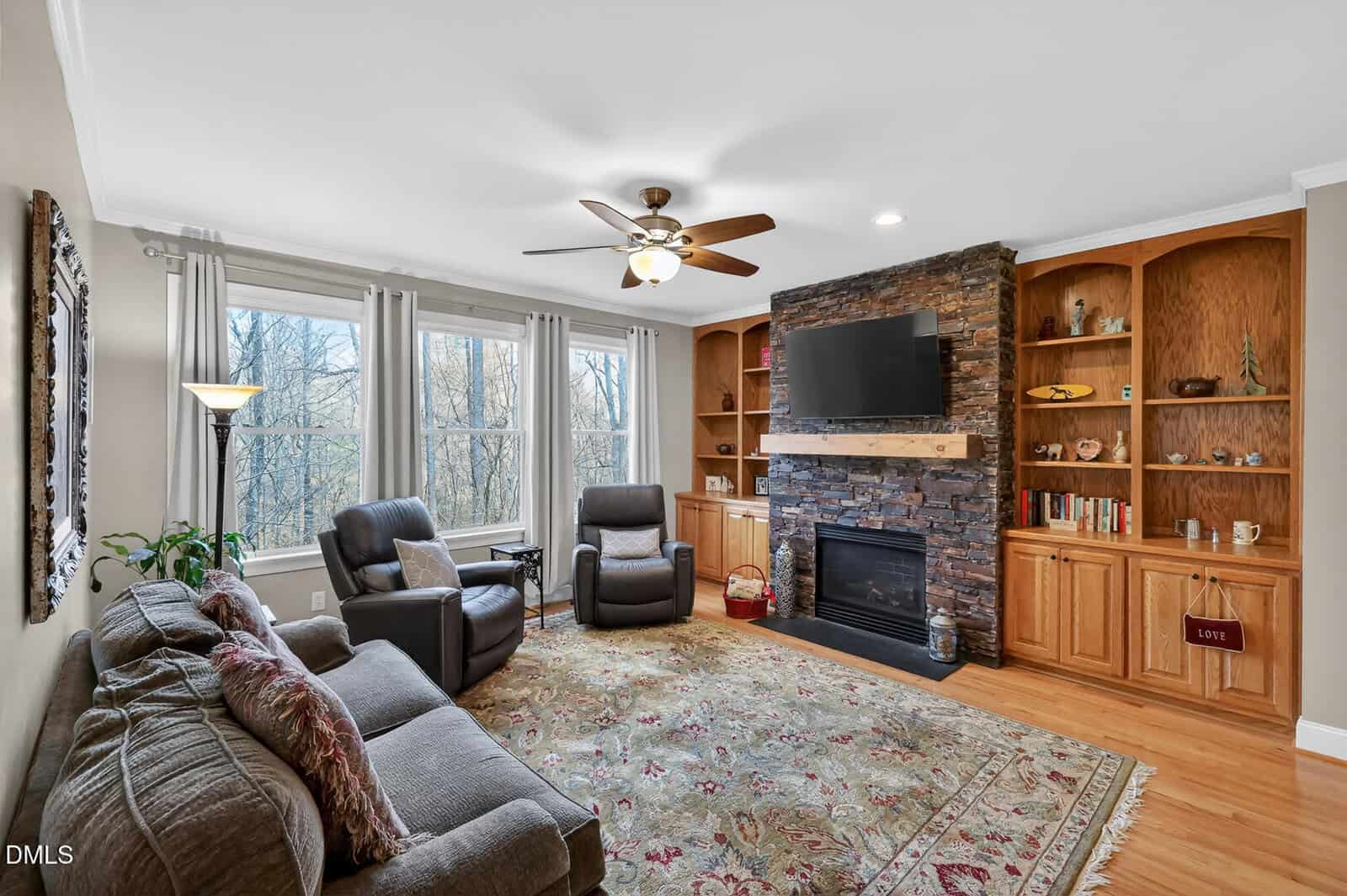 Living room with large windows, a ceiling fan, a couch, two recliners, a fireplace with a stone surround, a wall-mounted TV, built-in wood shelves, and a patterned rug on hardwood floors.