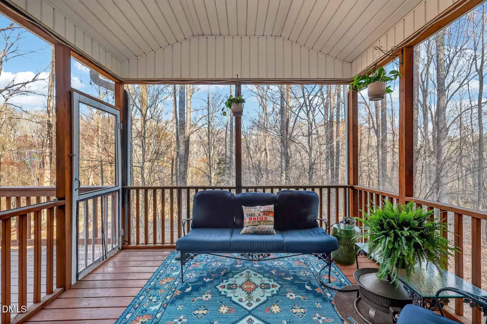 A screened-in porch with wooden flooring, a blue cushioned loveseat, a patterned rug, side tables with potted plants, and hanging planters. Leafless trees are visible through the screen, suggesting late fall or winter.