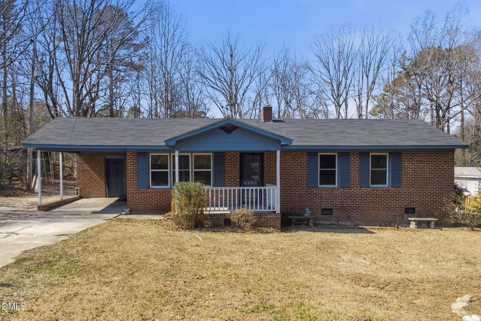 A single-story brick house with a dark roof, blue trim, and a small front porch sits on a patchy, dry lawn. Leafless trees fill the background under a clear sky. A driveway runs along the left side of the house.