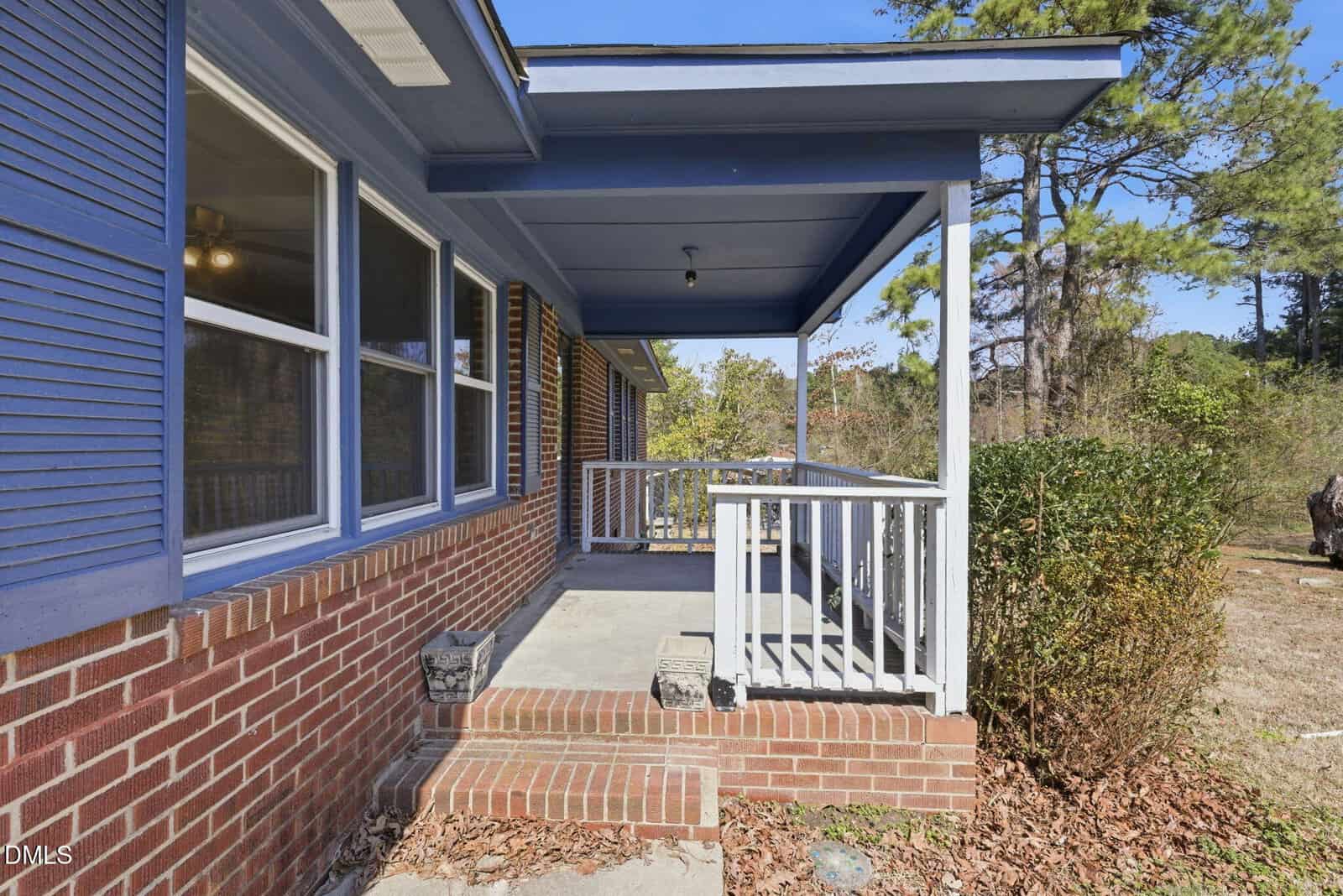 A brick porch with three steps leading to a covered entryway. The porch has white railings, blue trim, and three windows on the left. Bushes and trees are visible in the background under a clear sky.