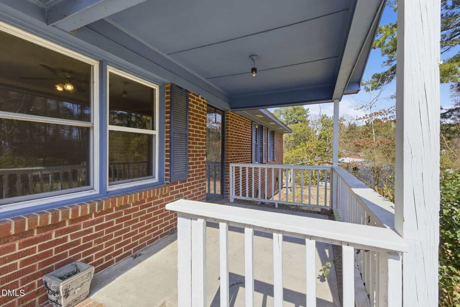 A covered front porch with a red brick exterior, blue trim, white wooden railing, and two large windows; a small planter sits on the concrete floor near the corner. Trees and shrubs are visible in the background.