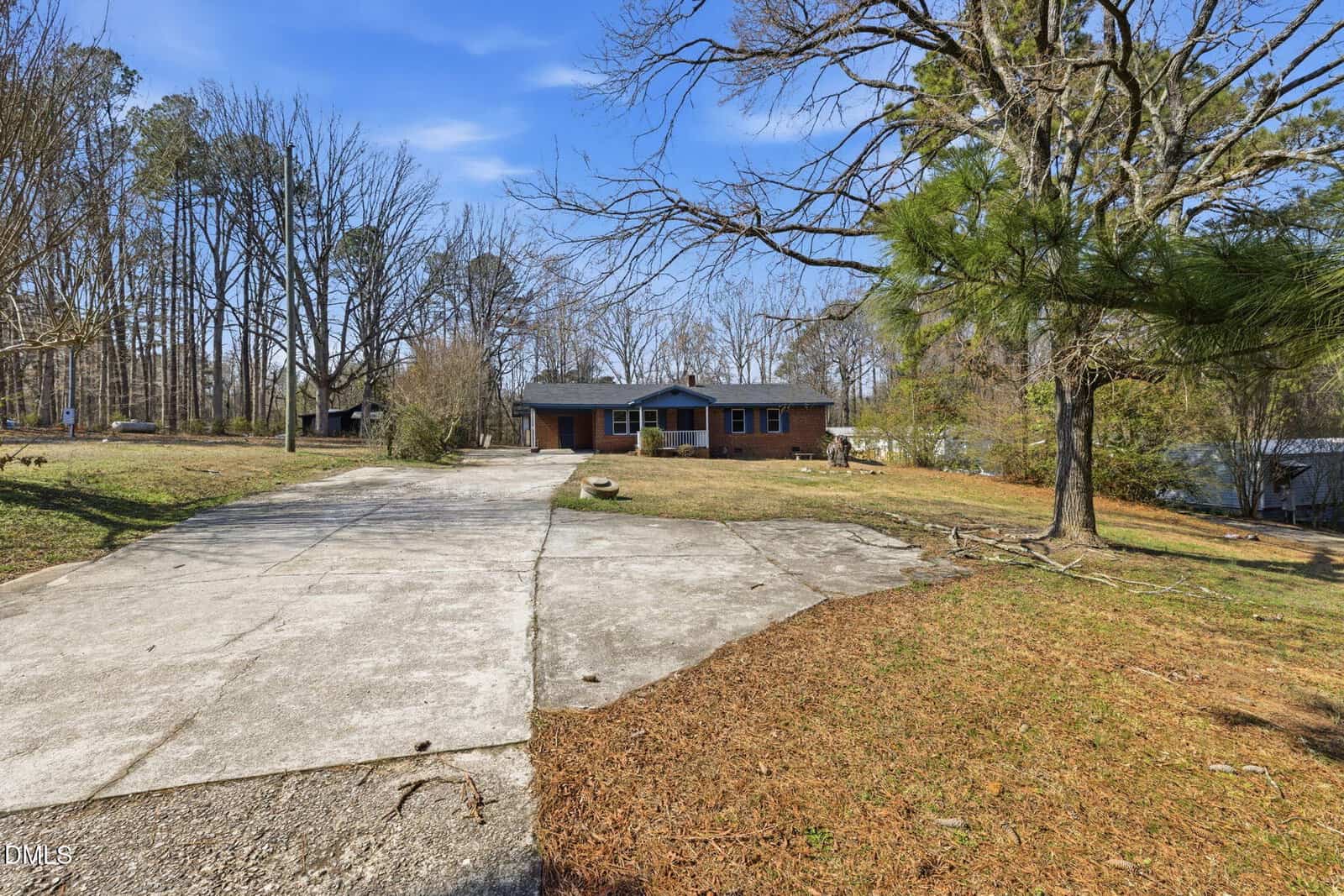 A single-story brick house sits at the end of a wide, cracked concrete driveway. The front yard has sparse grass, scattered leaves, and mature trees. The sky is clear and blue.