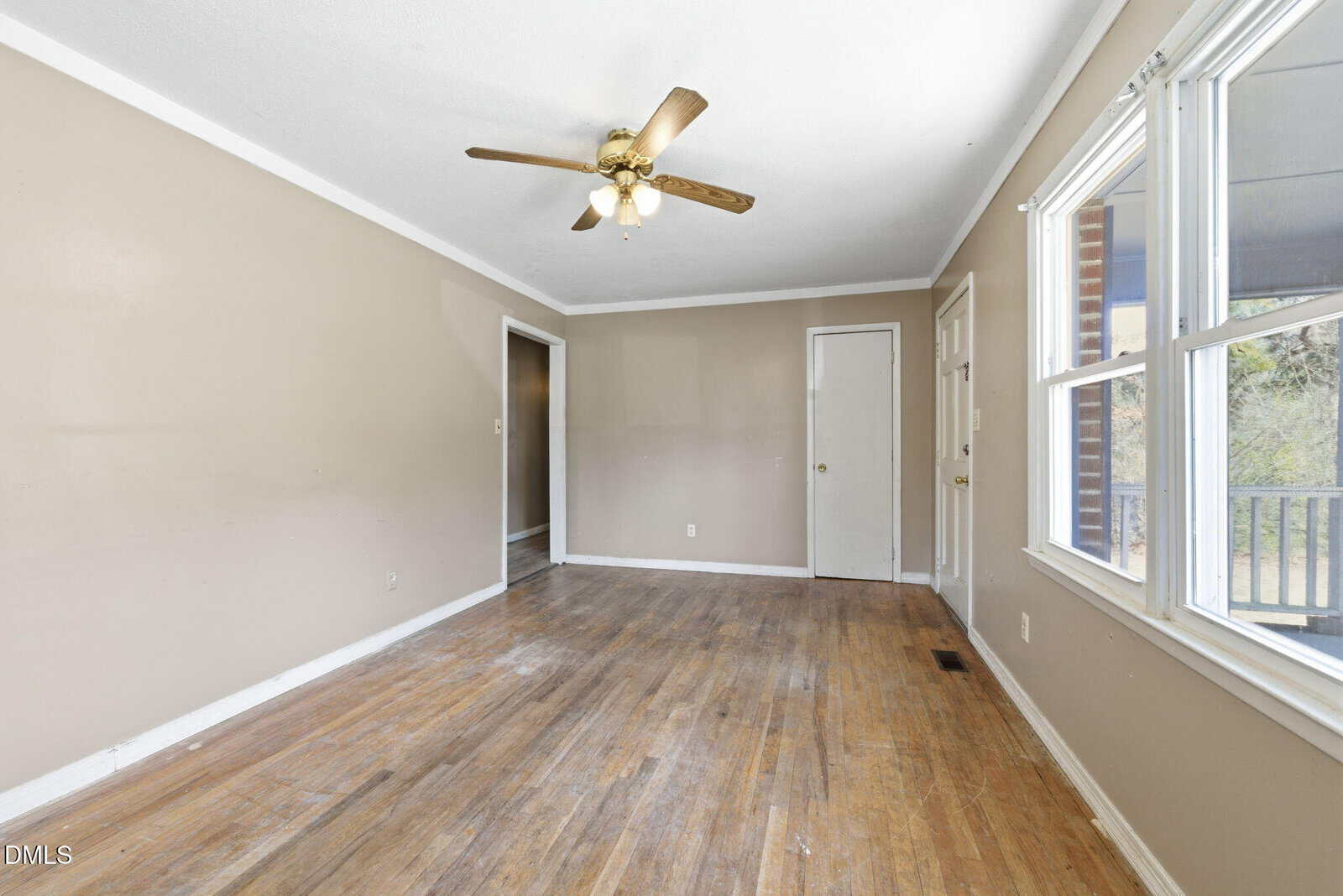 Empty room with beige walls, a white ceiling with a ceiling fan, two windows on the right letting in sunlight, worn hardwood floors, and a door and doorway visible in the background.