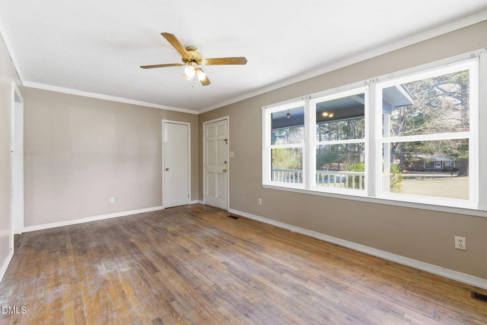 Empty room with wood floors, beige walls, a ceiling fan with lights, and three large windows letting in natural light. The room also has a white door and minimal trim. Some floor areas show noticeable wear.