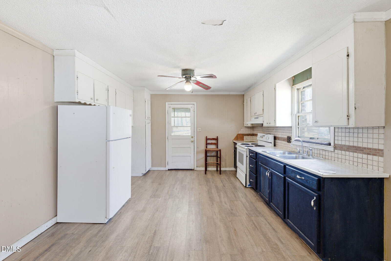 A kitchen with light wood floors, white upper cabinets, dark blue lower cabinets, a white refrigerator, stove, and dishwasher. A ceiling fan is centered above, and a single chair sits by the back door.