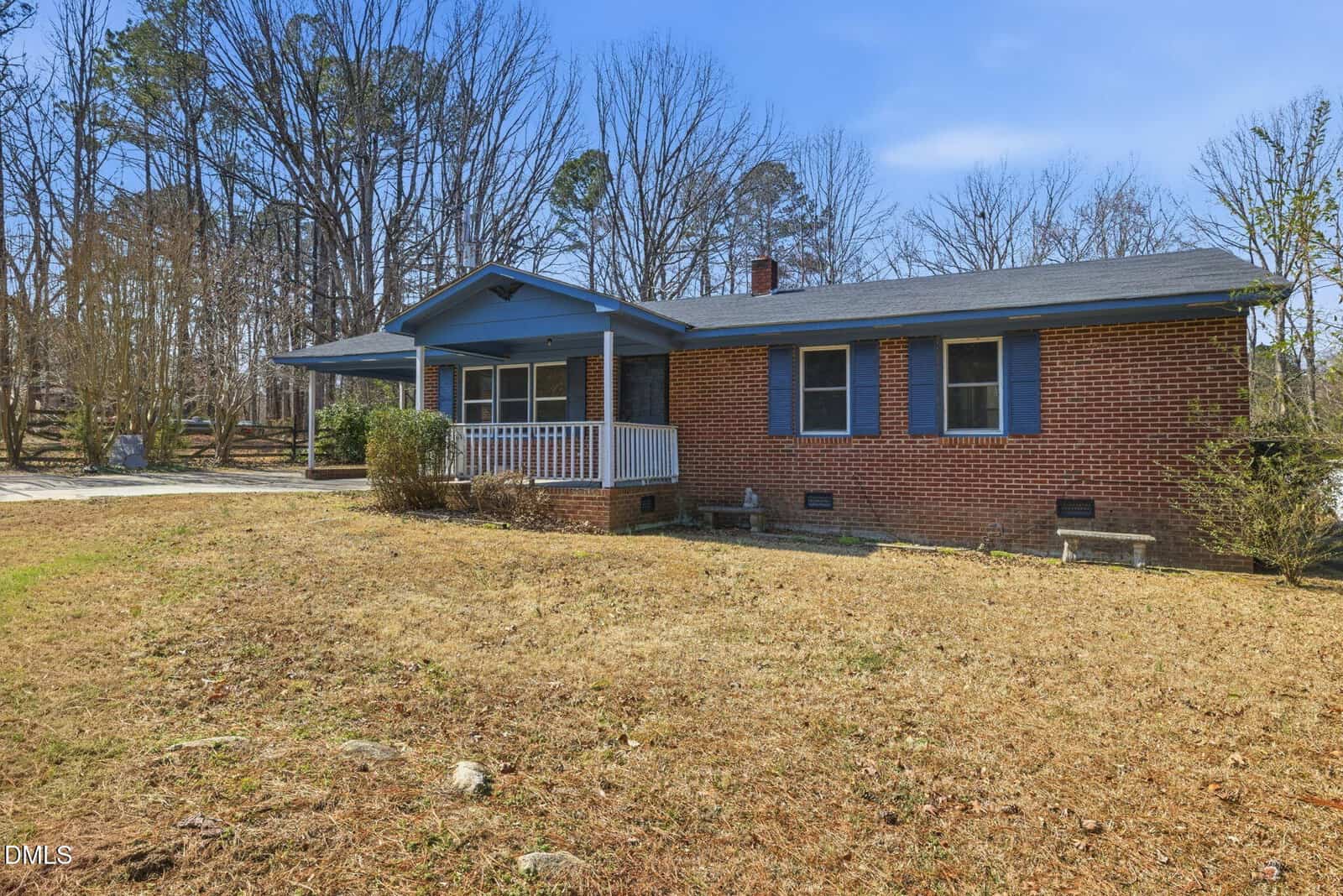 A single-story brick house with a covered front porch, blue trim, and multiple windows sits on a grassy yard with bare trees in the background under a clear sky.
