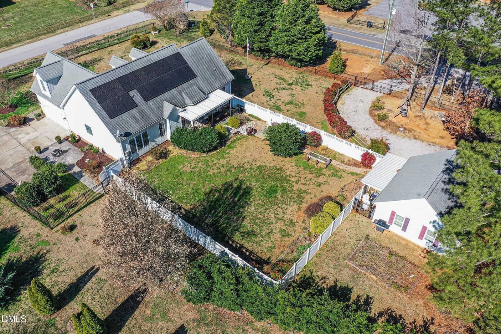 Aerial view of a white house with solar panels on the roof, an attached garage, a fenced backyard with patchy grass and shrubs, a detached outbuilding, and surrounding trees near a road.