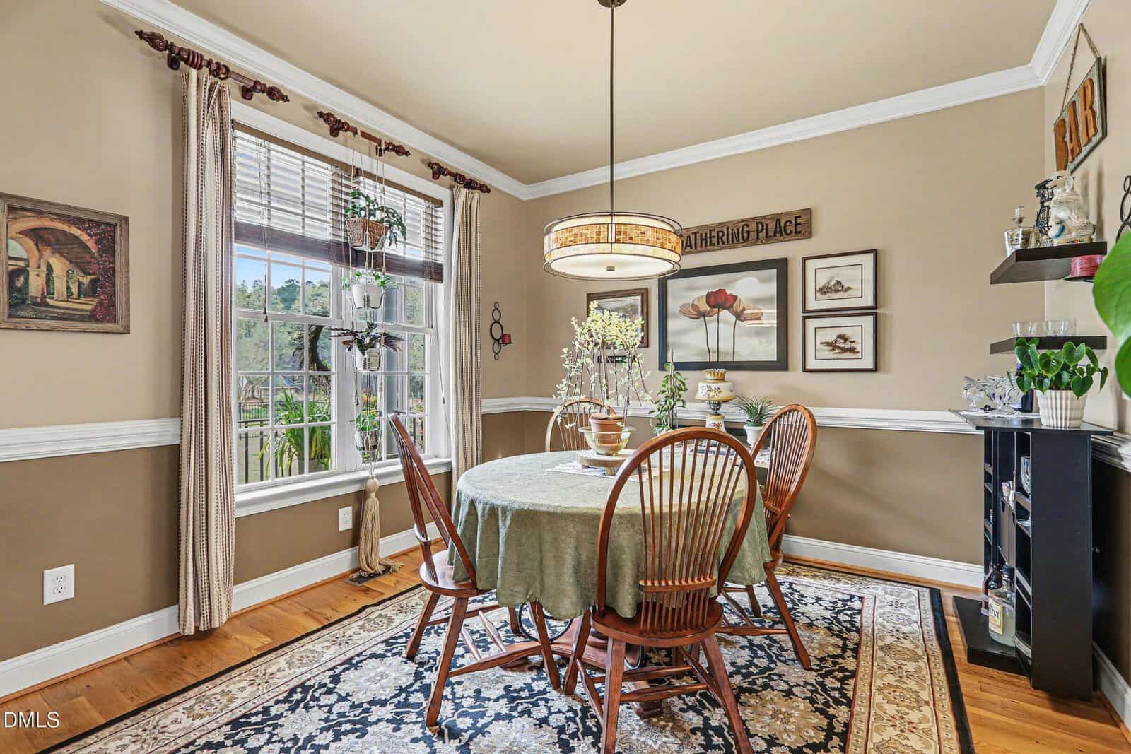 A dining room with tan walls, a round table covered with a green cloth, four wooden chairs, and a large window with sheer curtains. A decorative rug and various wall art and plants are also visible in the space.