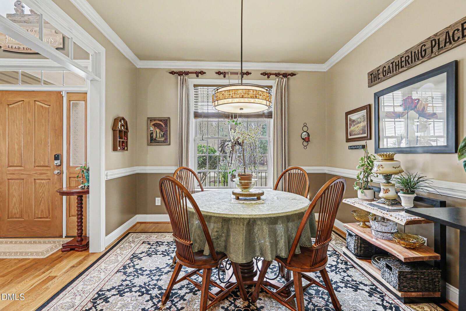 A dining room with a round table covered by a tablecloth, four wooden chairs, a hanging light fixture, a window with blinds, a front door, and shelves with plants and decorative items on the right wall.