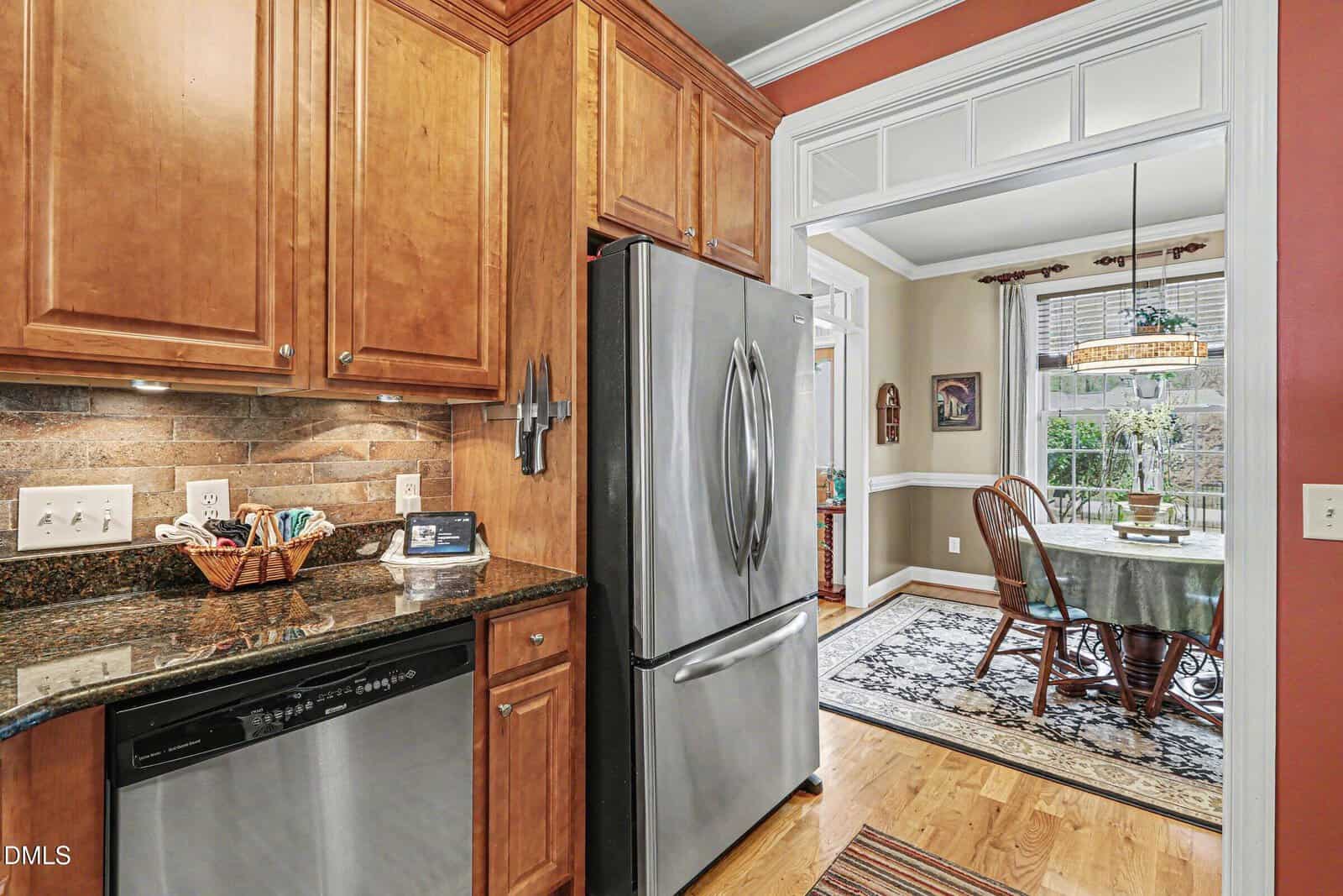 A kitchen with wooden cabinets, granite countertops, stainless steel dishwasher and refrigerator, and a view into a dining room with a table, chairs, rug, and large window.