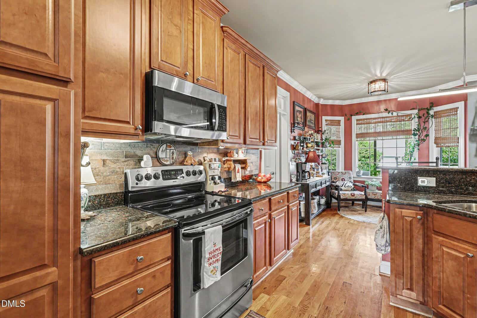 A kitchen with wooden cabinets, stainless steel appliances, granite countertops, and a hardwood floor. The room extends into a sitting area with windows, a round rug, and decorative items on shelves.