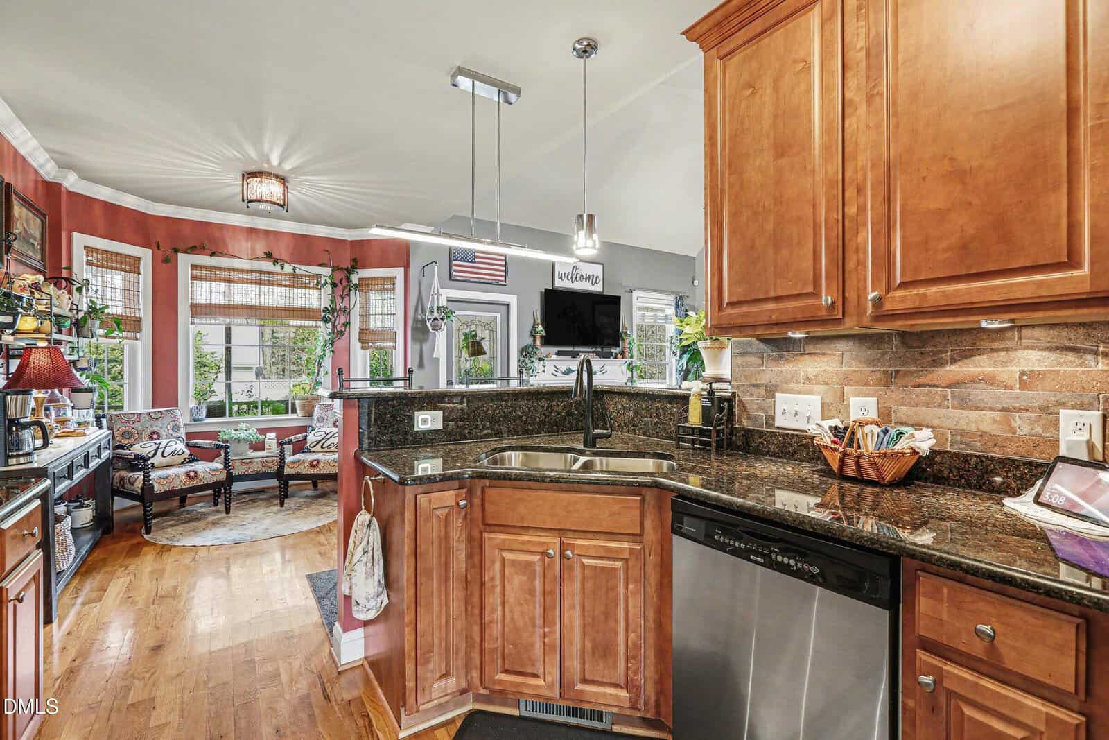A kitchen with wooden cabinets, granite countertops, brick-style backsplash, stainless steel dishwasher, and a double sink, opening into a bright sitting area with plants and large windows.