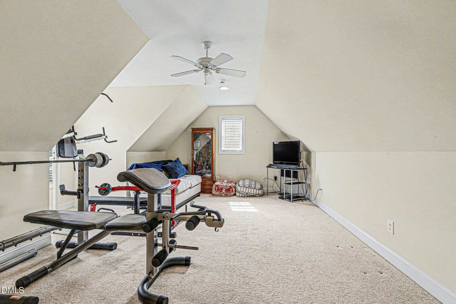 A carpeted attic room with angled ceilings contains a weight bench, a bed with blue bedding, a mirror, a TV on a stand, a ceiling fan, and a window letting in natural light.