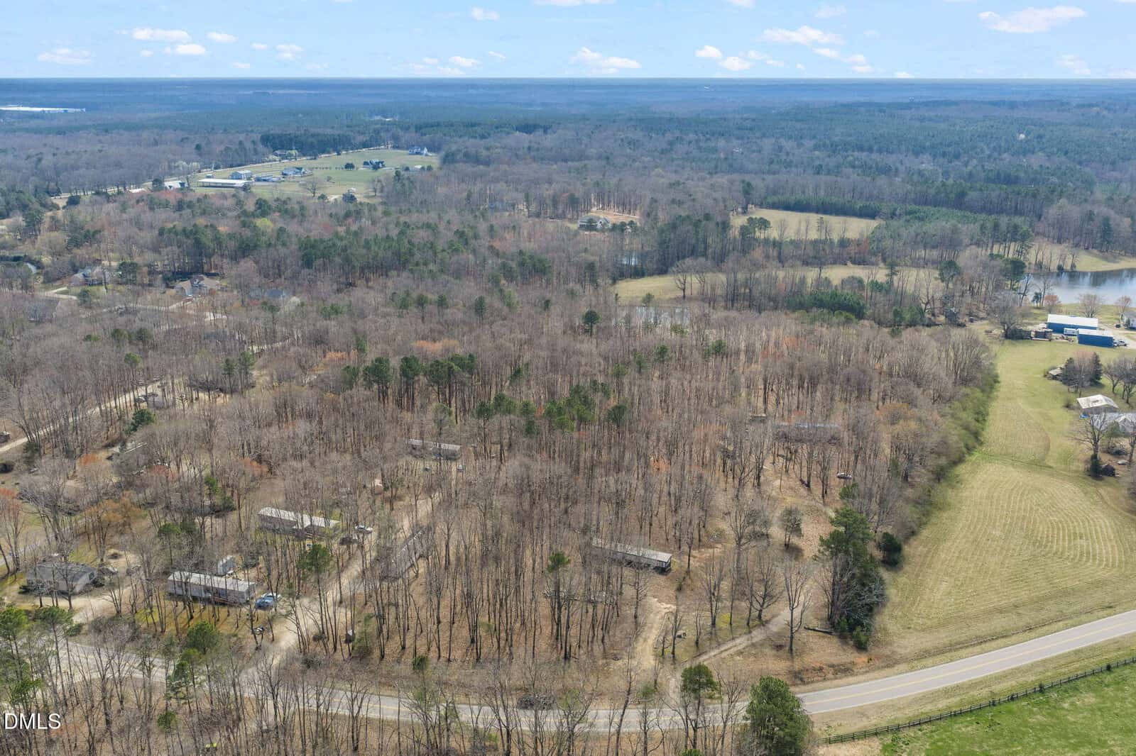 Aerial view of a rural landscape with scattered houses among leafless trees, open grassy areas, and a curving road in the foreground near 4074 Graham Sherron Road, with a distant forested horizon under a partly cloudy sky in Wake Forest.