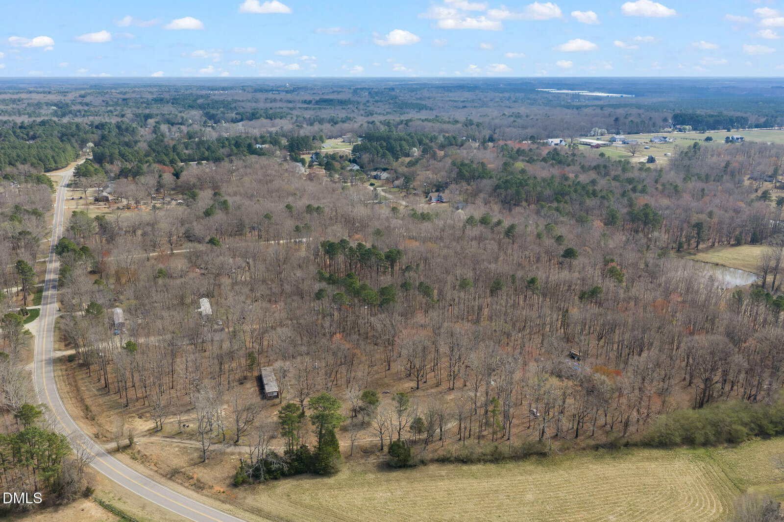 Aerial view of a rural landscape with leafless trees, winding roads, scattered houses, open fields, and distant forests under a partly cloudy sky.