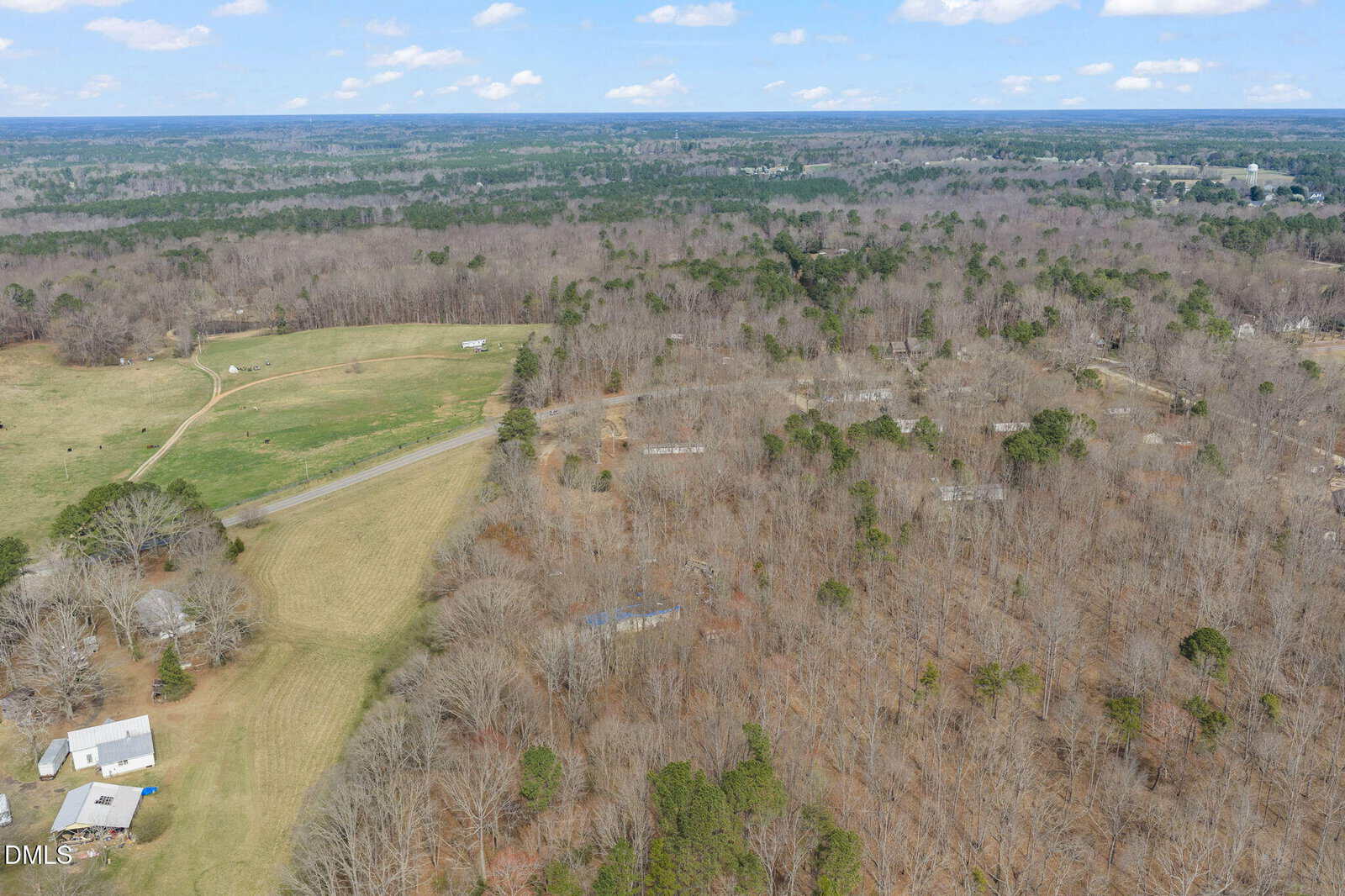Aerial view of a rural landscape with open fields, scattered trees, several buildings, and a road running through wooded and cleared areas under a partly cloudy sky.