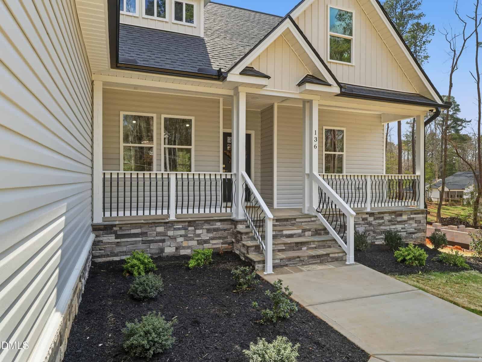 A modern house with light-colored siding, stone accents, a covered front porch with white railings, several steps leading up, and neatly landscaped shrubs along a concrete walkway.