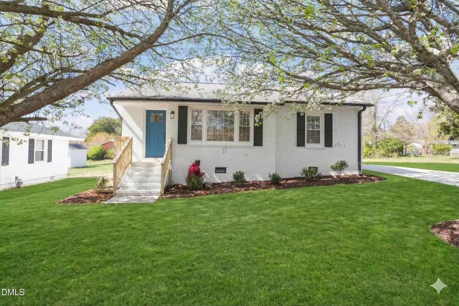 A single-story white brick house at 116 Halifax Street, Oxford, features a blue front door, small front porch with steps, large front windows, and manicured lawn. A person is kneeling near shrubs under a tree with budding leaves.