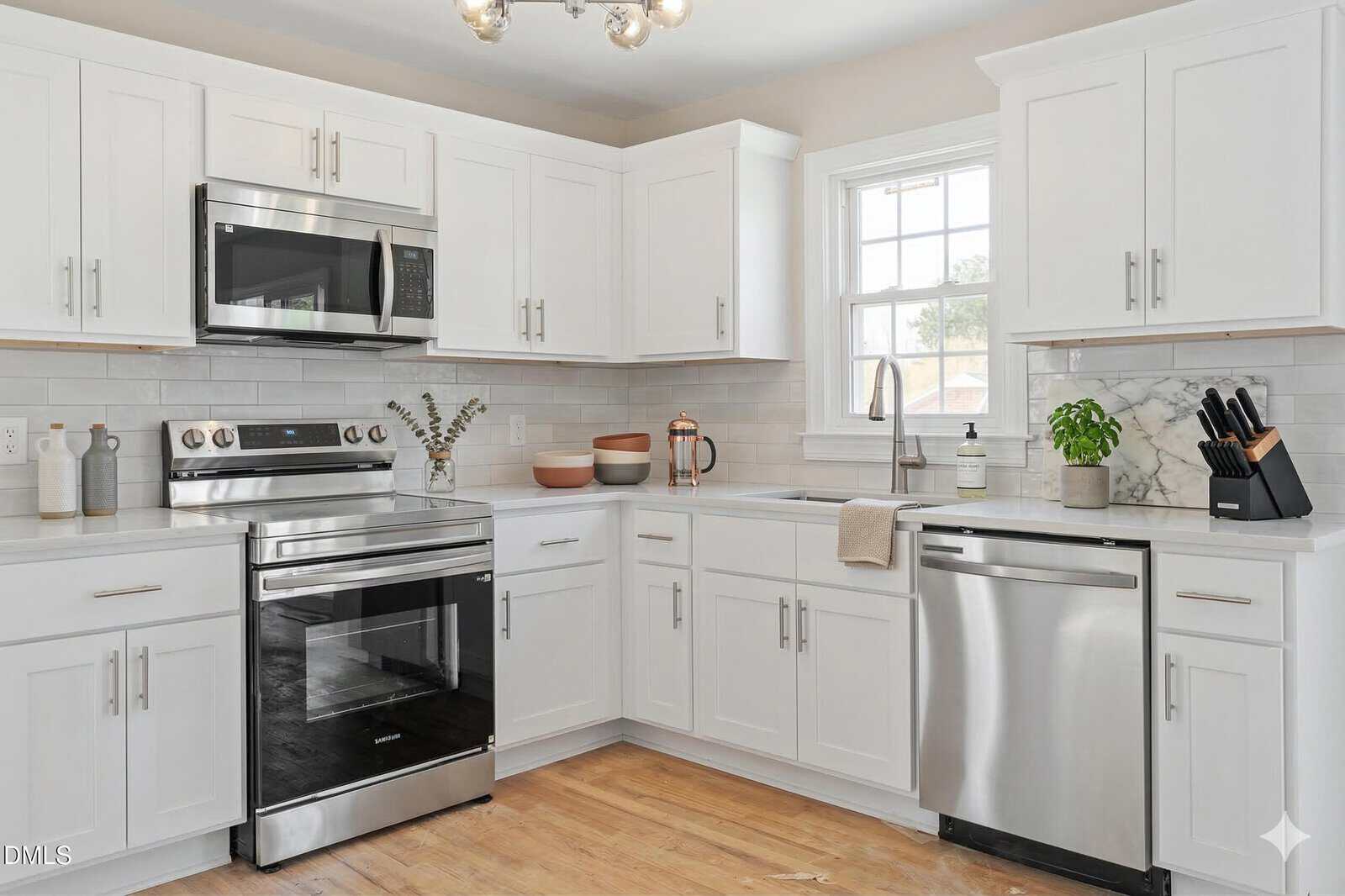 Modern kitchen with white cabinets, stainless steel appliances, a window above the sink, light wood flooring, a dishwasher, and minimalist decor including a small plant and various kitchenware on the counters.