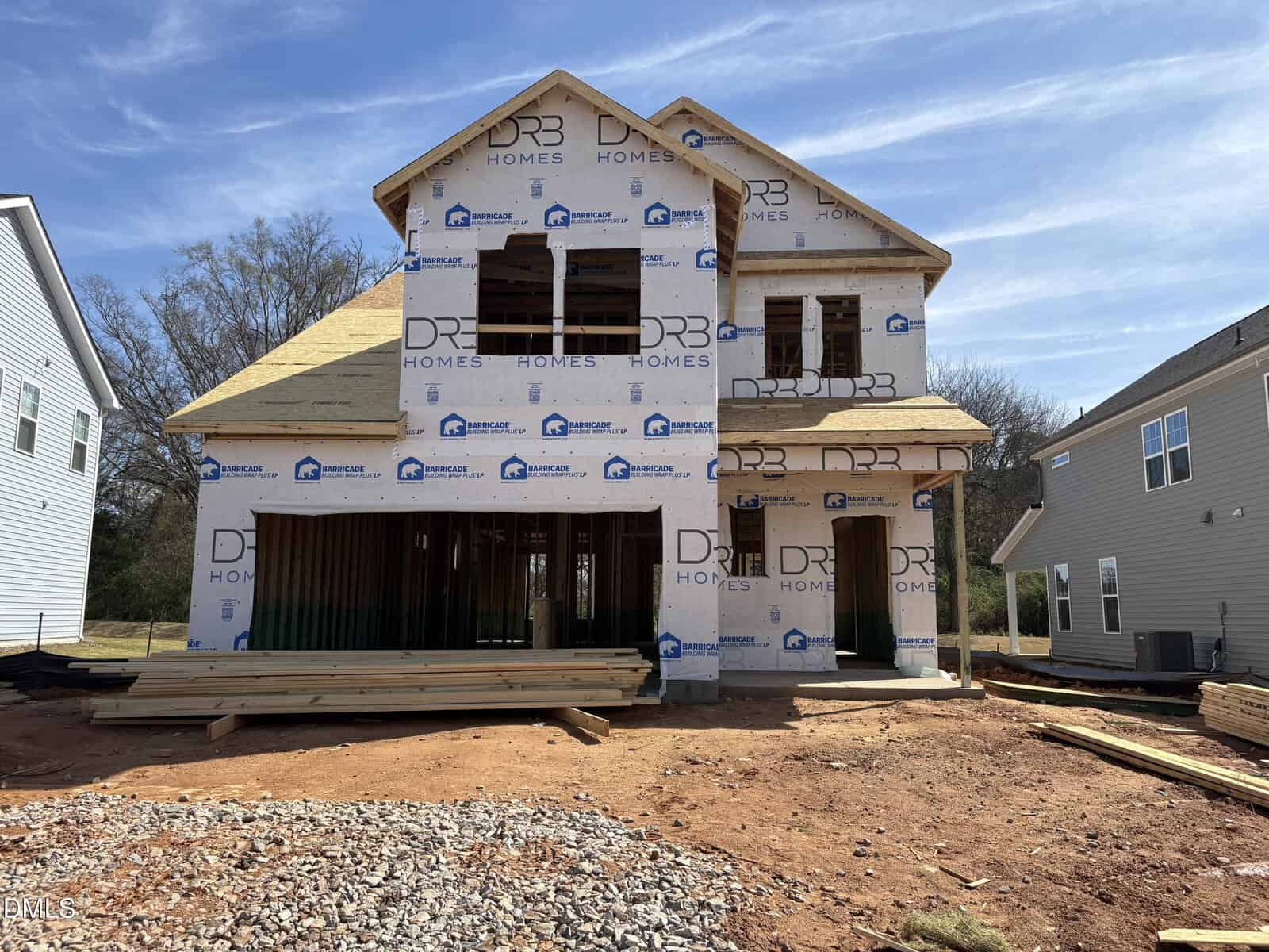 A two-story house under construction, covered with DRB Homes wrap. The structure has visible wood framing, open window spaces, and building materials stacked in the front yard. Dirt and gravel cover the ground.