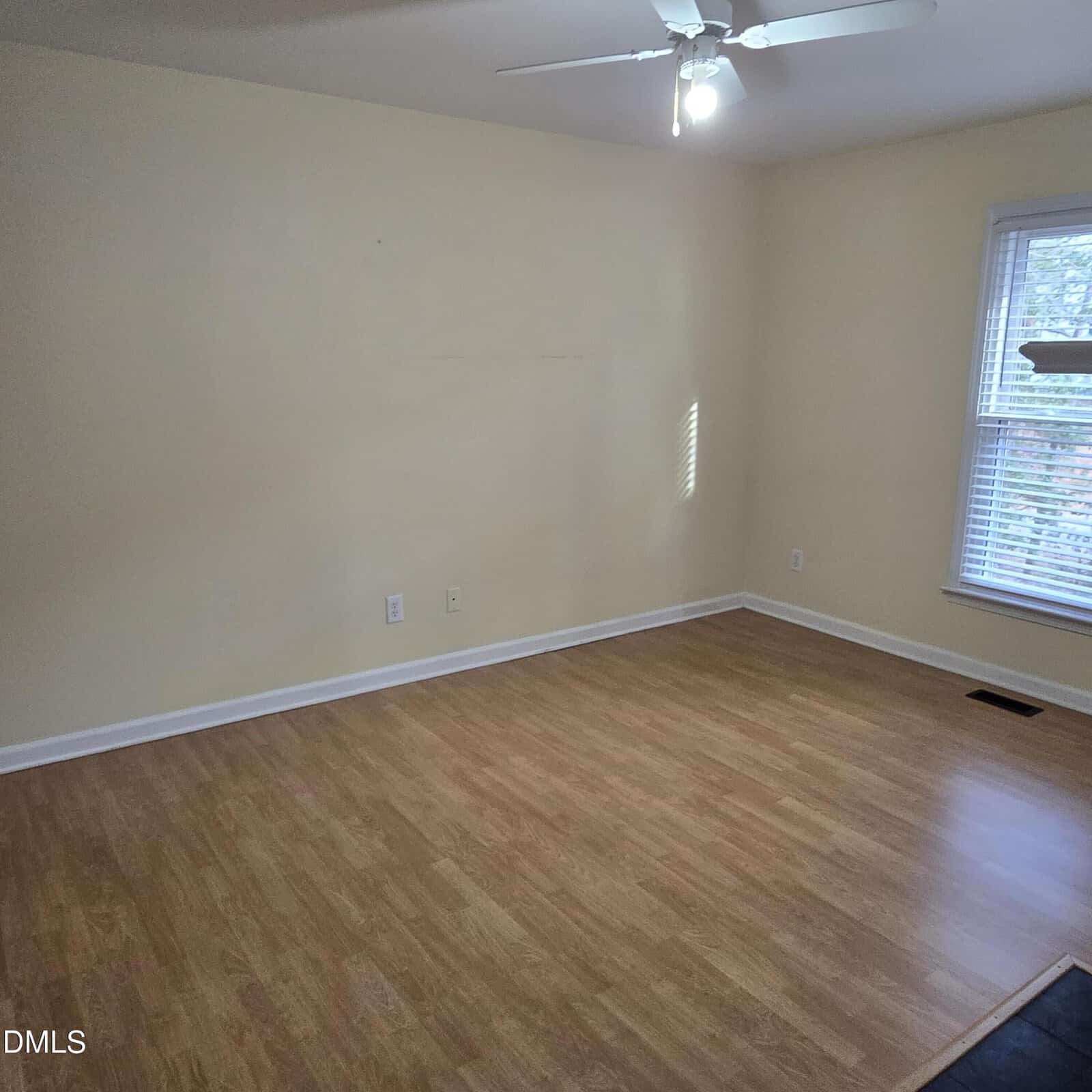 Empty room with light-colored walls, wood flooring, a ceiling fan with a light turned on, two electrical outlets on one wall, and a window with blinds letting in natural light.