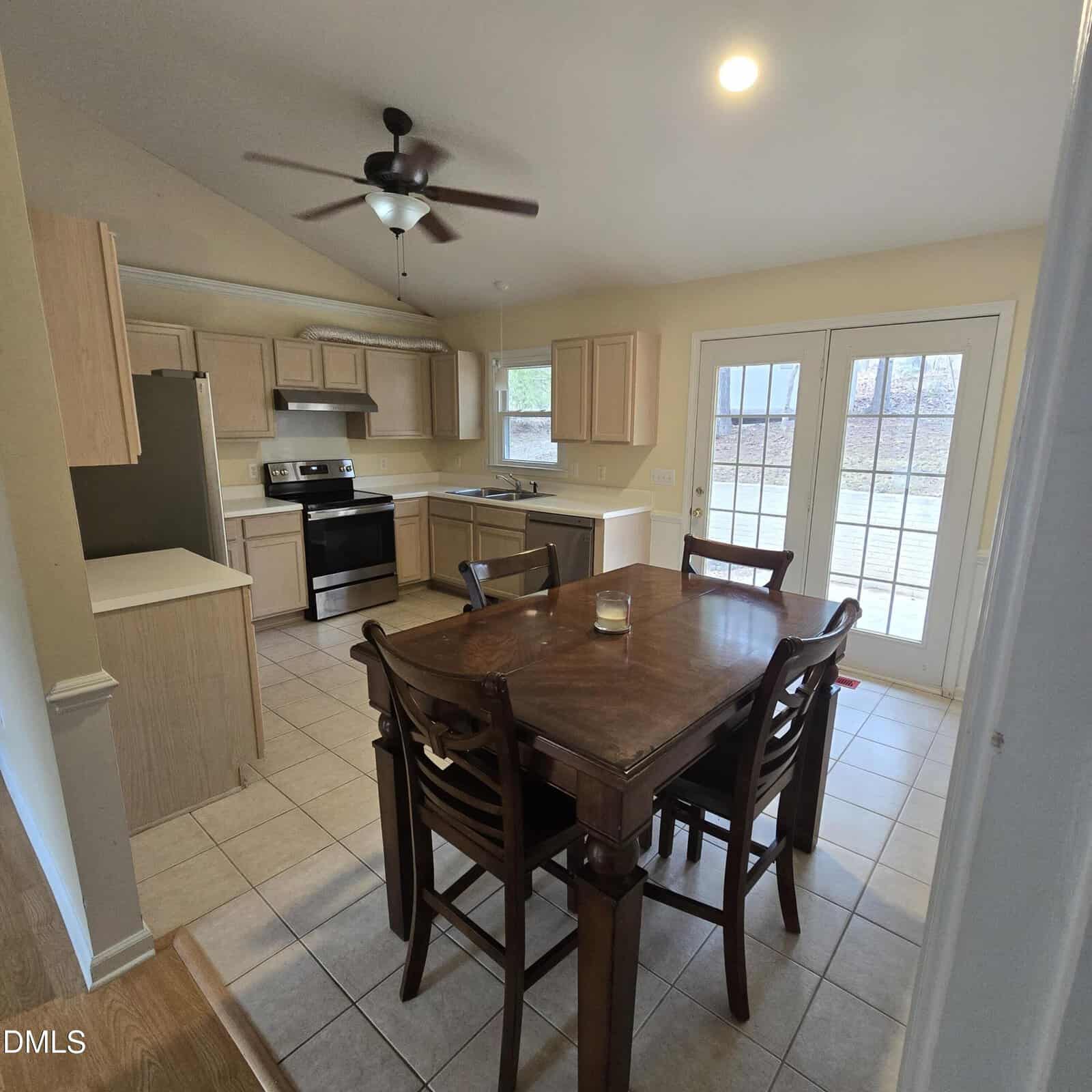 A kitchen and dining area with light wood cabinets, stainless steel appliances, a ceiling fan, a wooden dining table with four chairs, tiled flooring, and French doors leading outside.