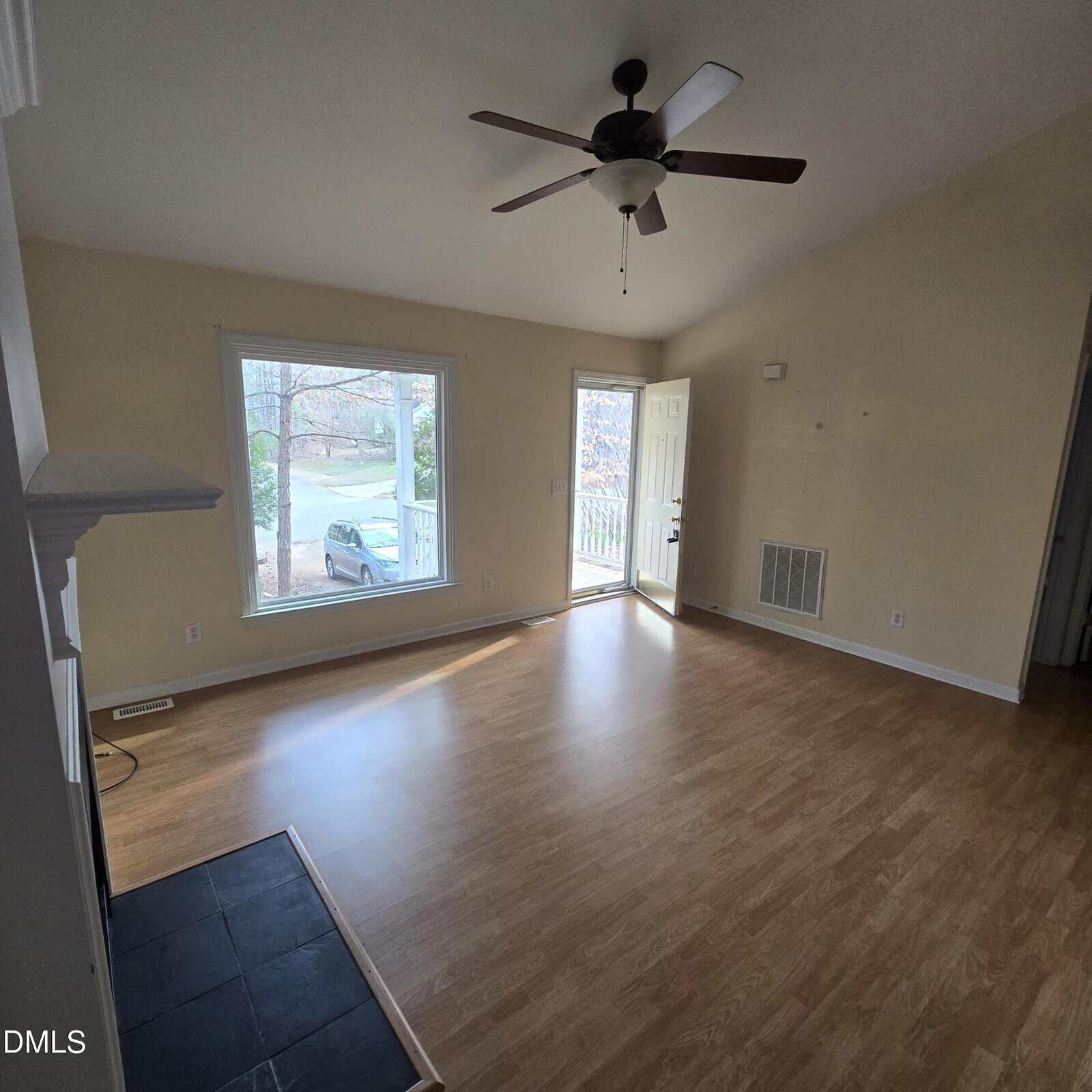 A bright, empty living room with light wood floors, large front window, ceiling fan, and front door open to the outside. Natural light fills the space, and a car is visible through the window.