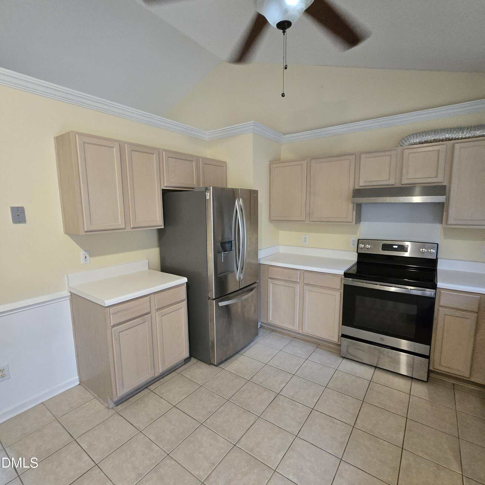A kitchen with light wood cabinets, a stainless steel refrigerator, and a stainless steel electric stove with a range hood. The space has beige tiled floors, pale yellow walls, and white countertops.
