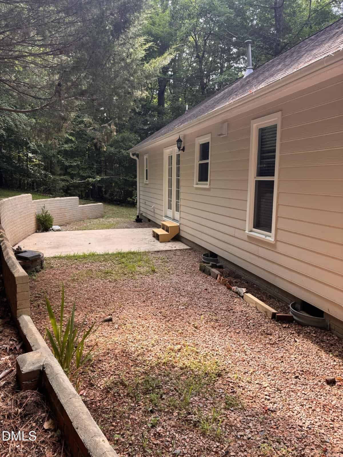 A beige house with white trim sits next to a gravel area and concrete patio, bordered by short brick retaining walls. Trees and dense woods are visible in the background. A small yellow step leads to a glass door.