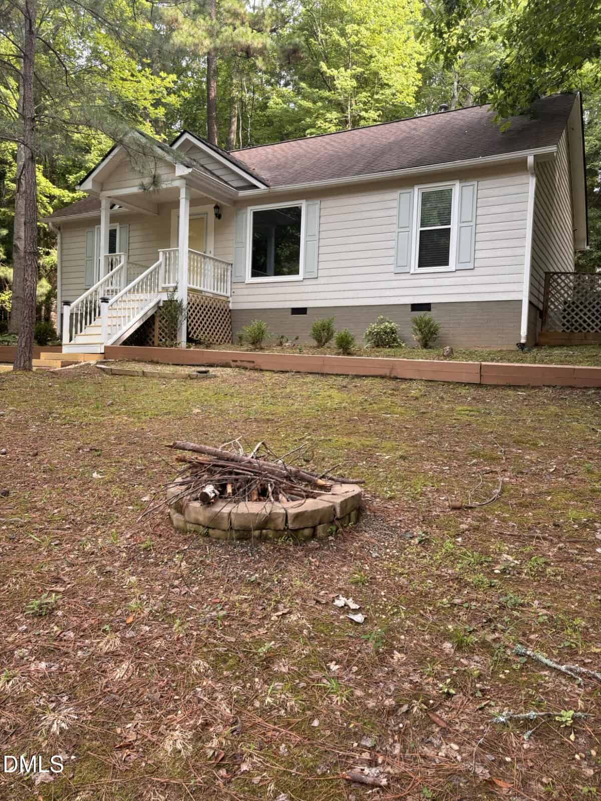 Single-story house with light-colored siding, a covered front porch, and white trim. The front yard has a small circular fire pit made of stones and a raised garden bed by the house, surrounded by trees.