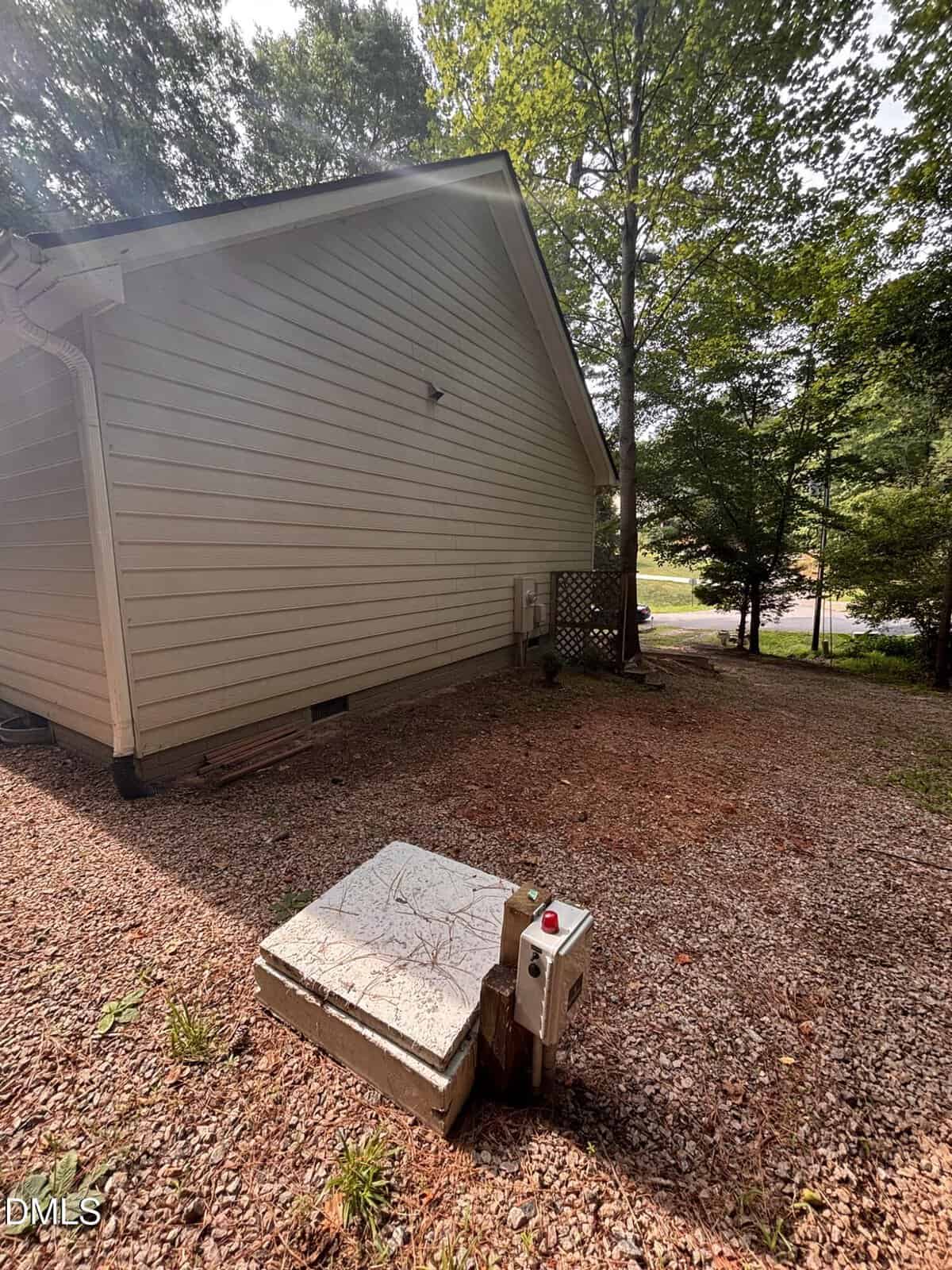 A concrete utility box with a metal control panel sits on gravel and dirt near the side of a light-colored house. Tall trees and a partially visible road are in the background.