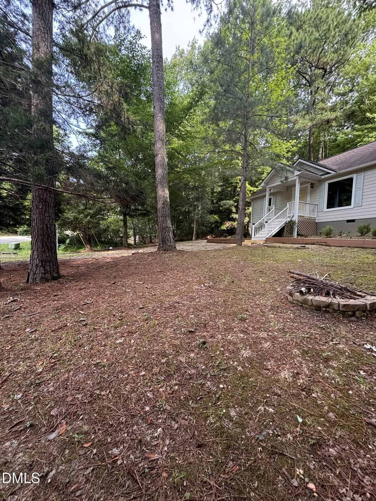 A grassy, pine needle-covered yard with tall trees, a small fire pit, and a white house with a porch and steps, set in a wooded area under a partly cloudy sky.