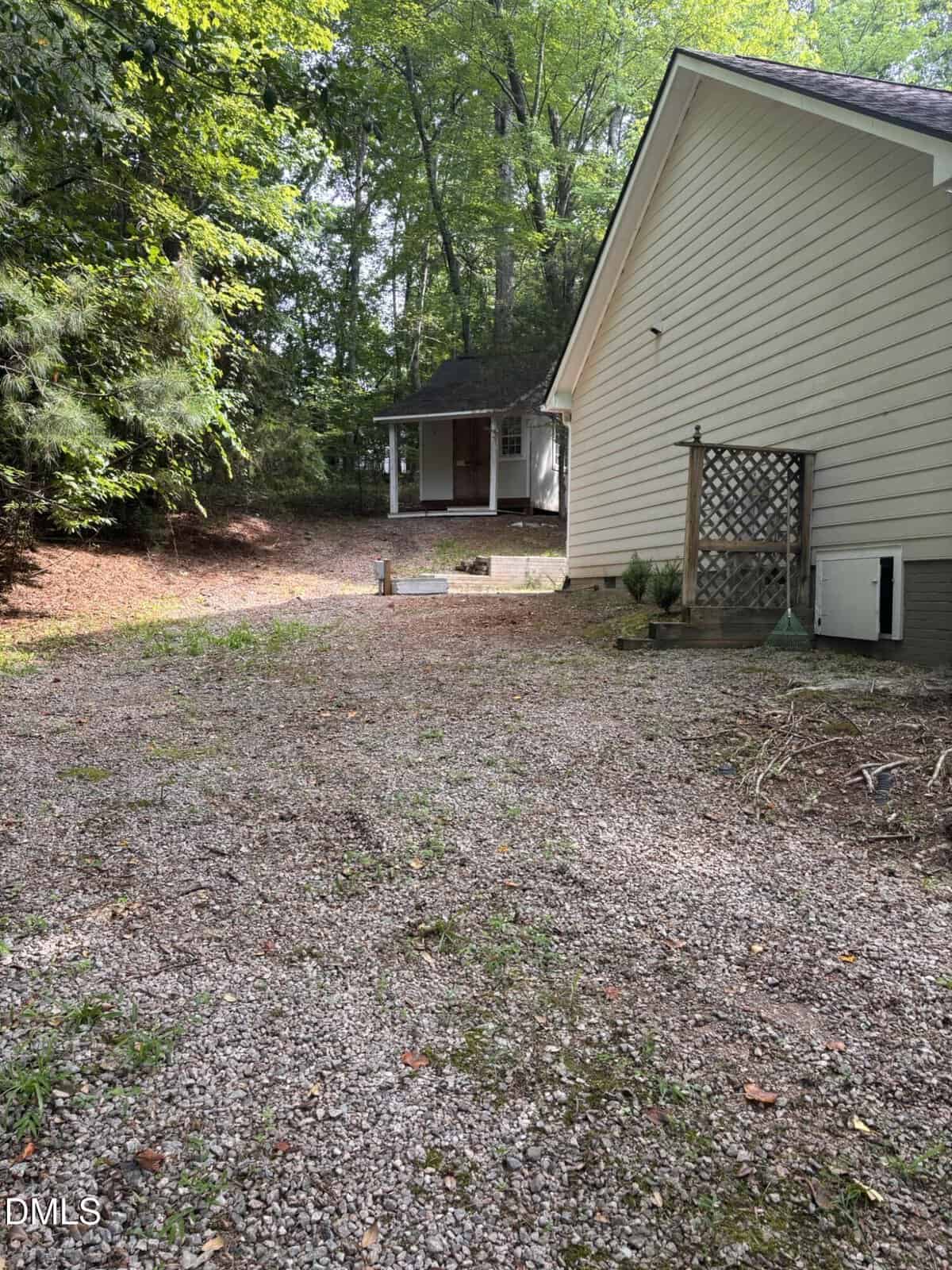 A gravel driveway leads to a small light-colored house with a porch, surrounded by trees and greenery. A wooden lattice and a few small plants are near the side of the house in the foreground.