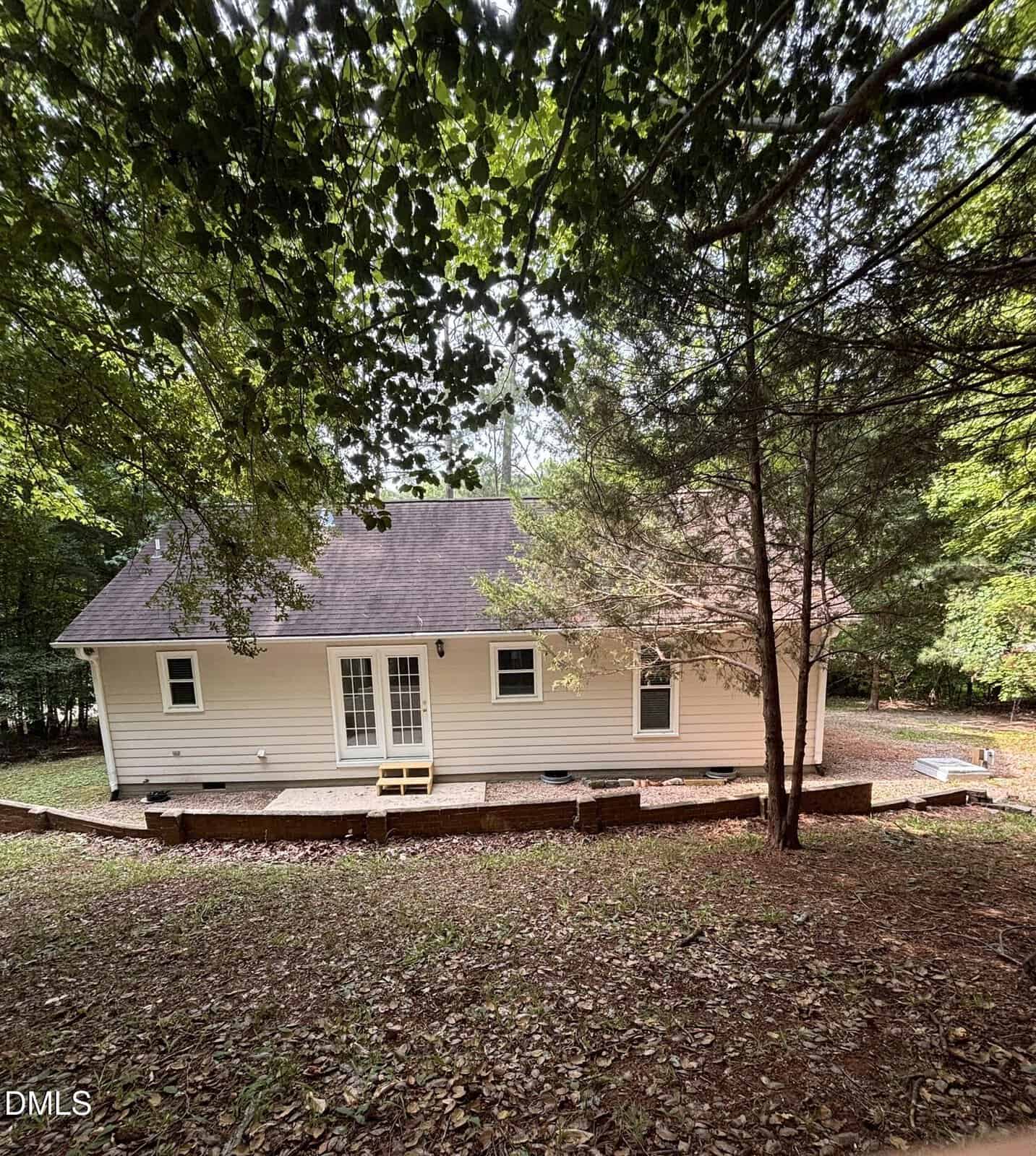 A small white house with a dark roof sits in a wooded area. There is a wooden deck at the back with steps leading down to a yard covered in dry leaves. Trees surround the house, providing shade.