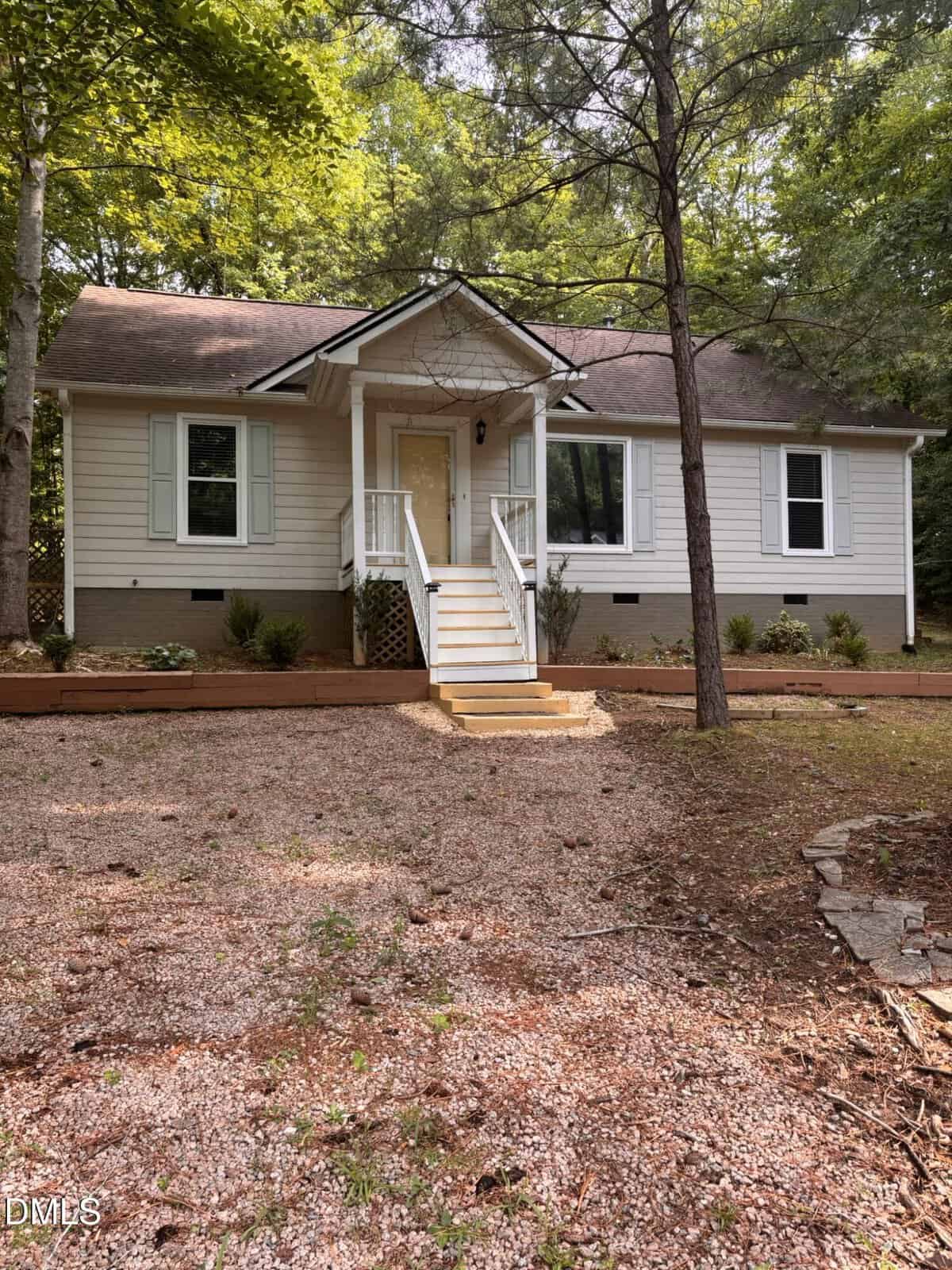 A small, light-colored house with white trim, shutters, and a front porch sits among tall trees at 113 Cochise Drive in Louisburg. A gravel path leads to the steps, with simple landscaping featuring shrubs and mulch beds in the yard.