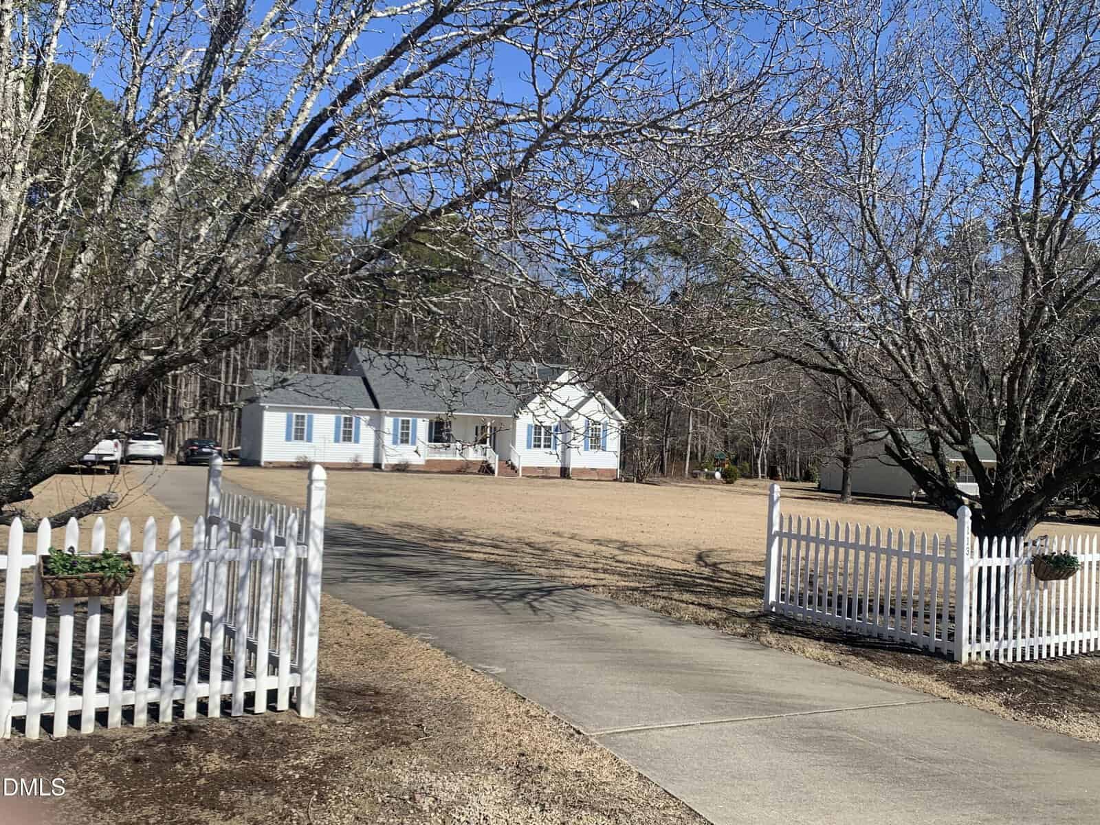 A white house with blue shutters sits at the end of a curved driveway, framed by leafless trees and a white picket fence on a sunny day. A detached garage is visible in the background, with a large yard in front.