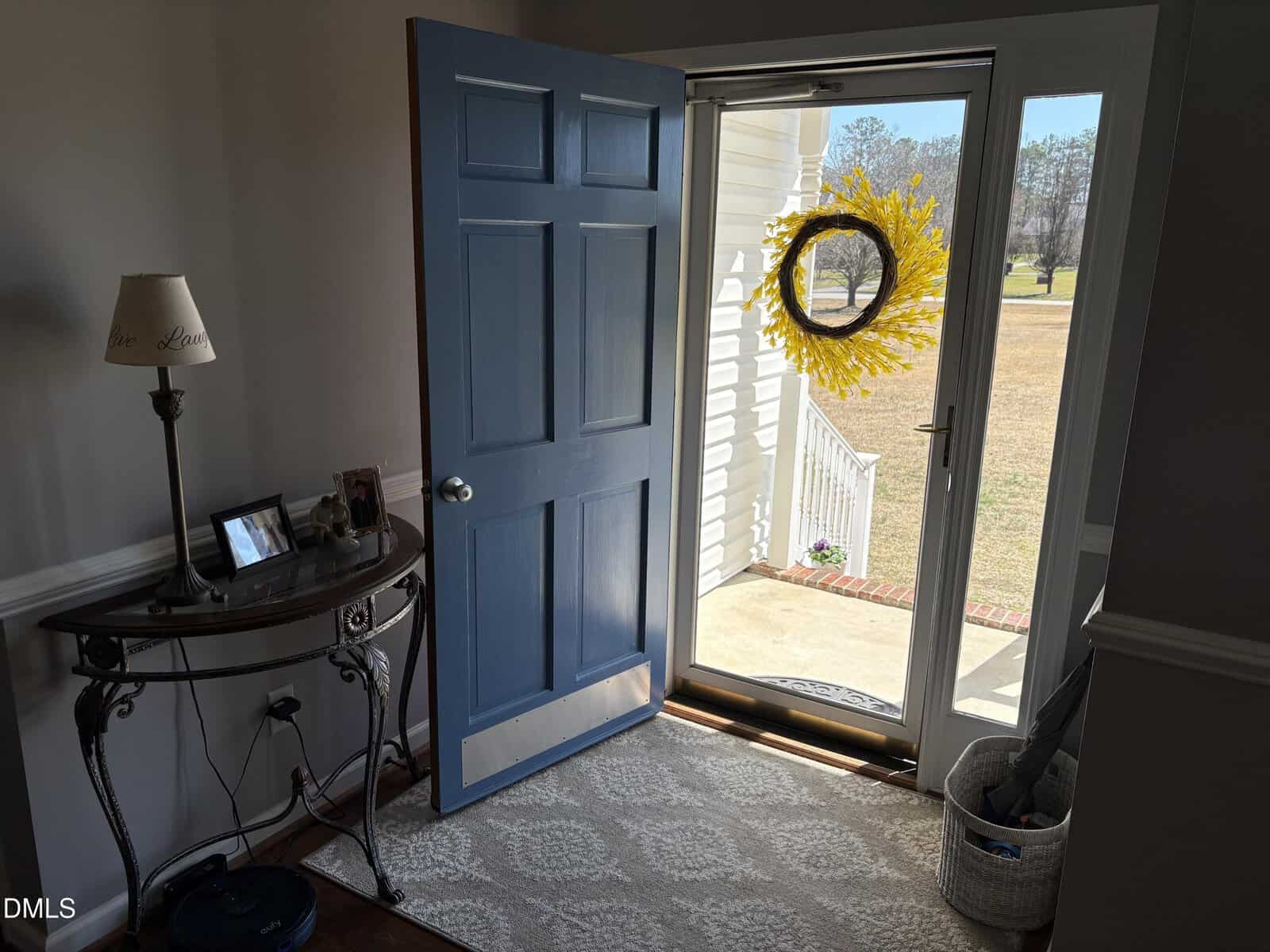 A sunlit entryway features a blue front door opened toward a glass storm door. A yellow wreath hangs on the outside door. Inside, a small table holds photos and a lamp, while a basket sits on a patterned rug by the doorway.