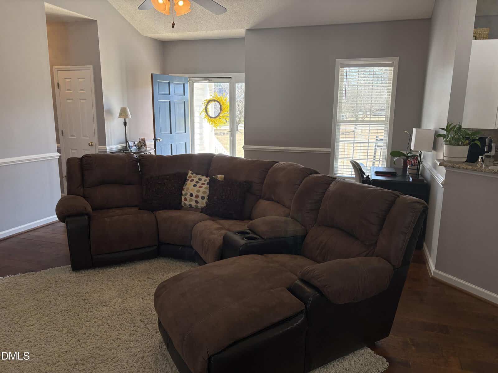 Living room with a brown sectional sofa on a light rug, a small desk by the window, open front door with a wreath, wood floor, light walls, and ceiling fan with lights. Natural light enters through windows.