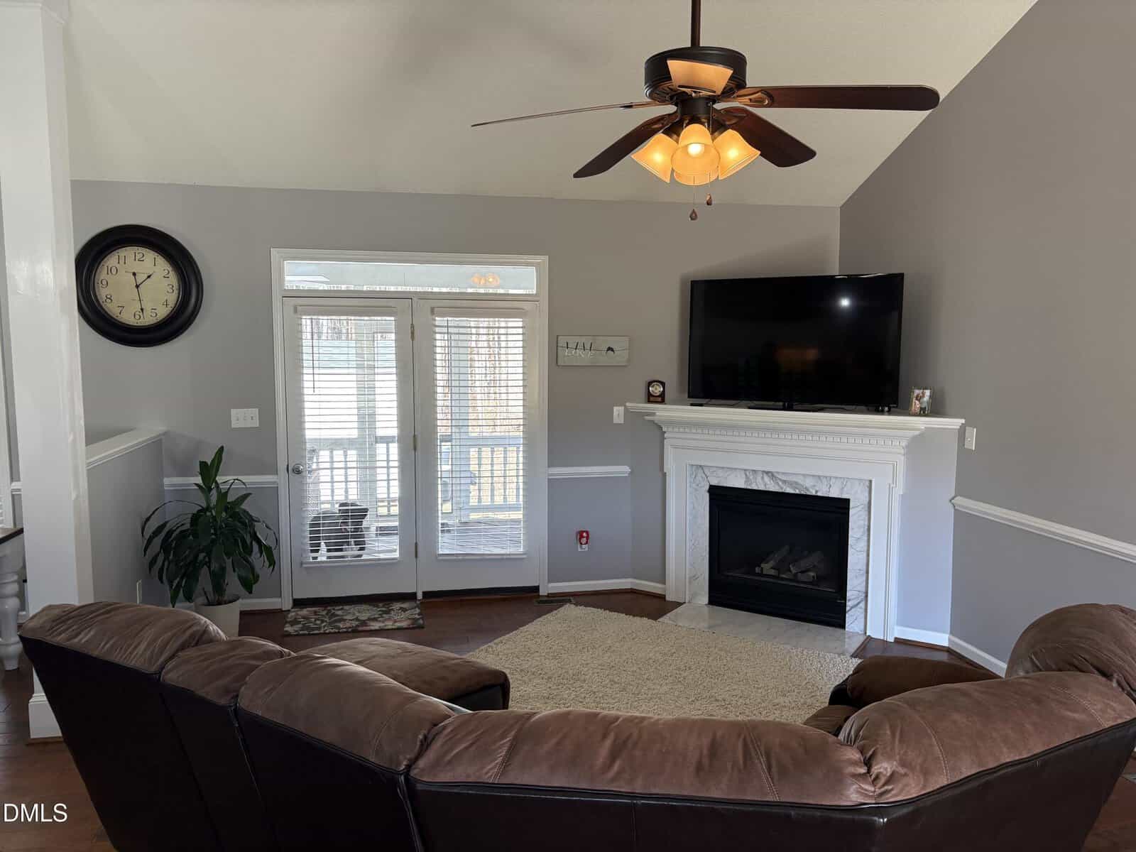 Living room with brown sectional sofa, area rug, wall-mounted TV above a white fireplace, clock on wall, ceiling fan with light, potted plant, and glass doors leading to a sunroom. Walls are painted light gray.