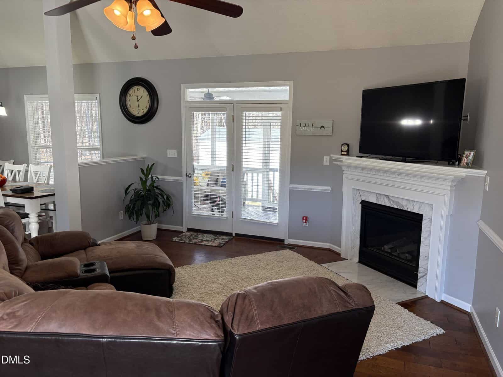 A living room with brown leather sofas, a fireplace with a TV above it, a wall clock, a potted plant, and glass doors leading outside. Wooden floors and neutral-colored walls complete the space.
