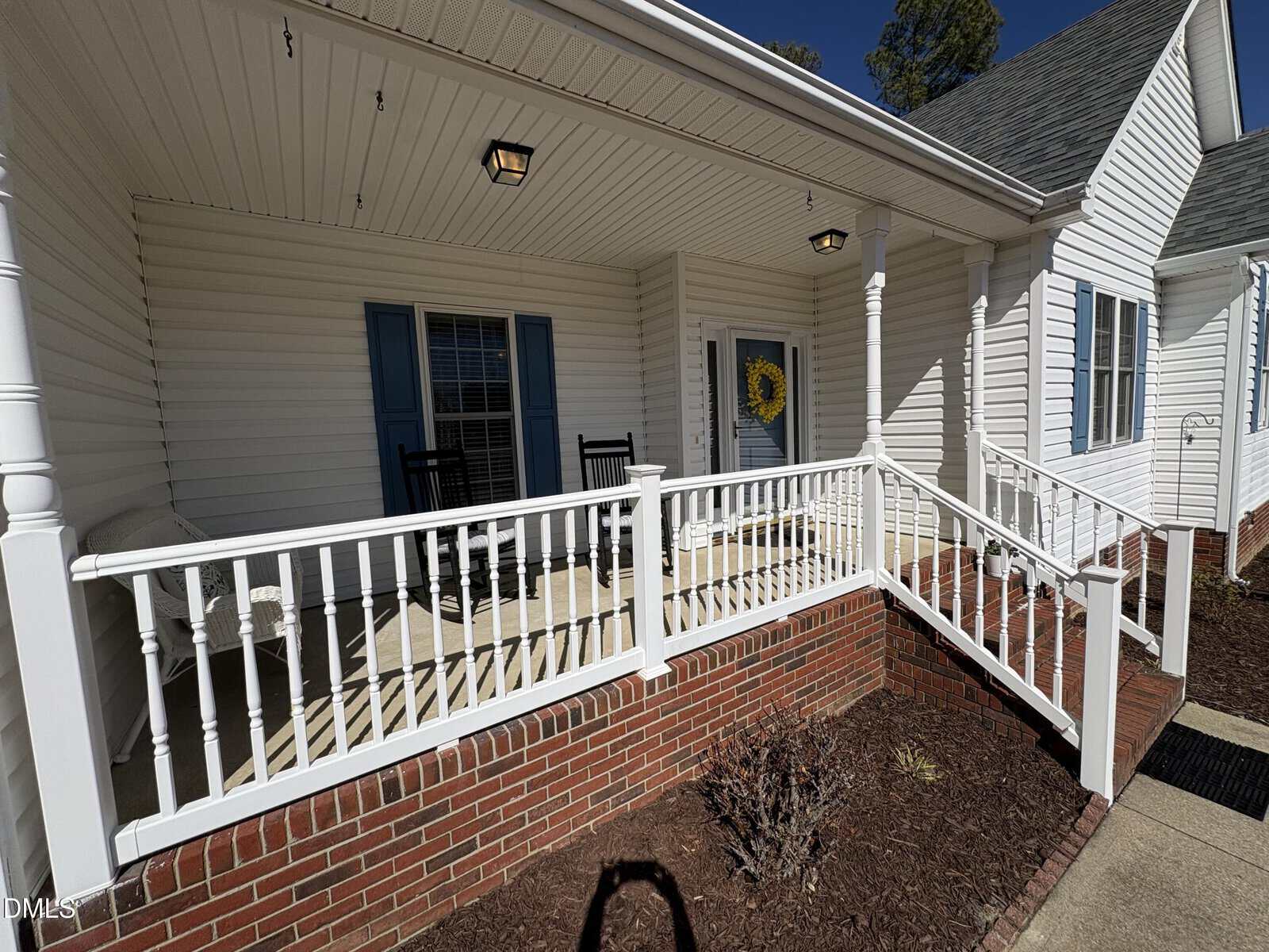 A front porch with white railings and columns, brick steps, two black rocking chairs, a bench, blue shutters, and a door decorated with a yellow wreath. The porch is attached to a white house.