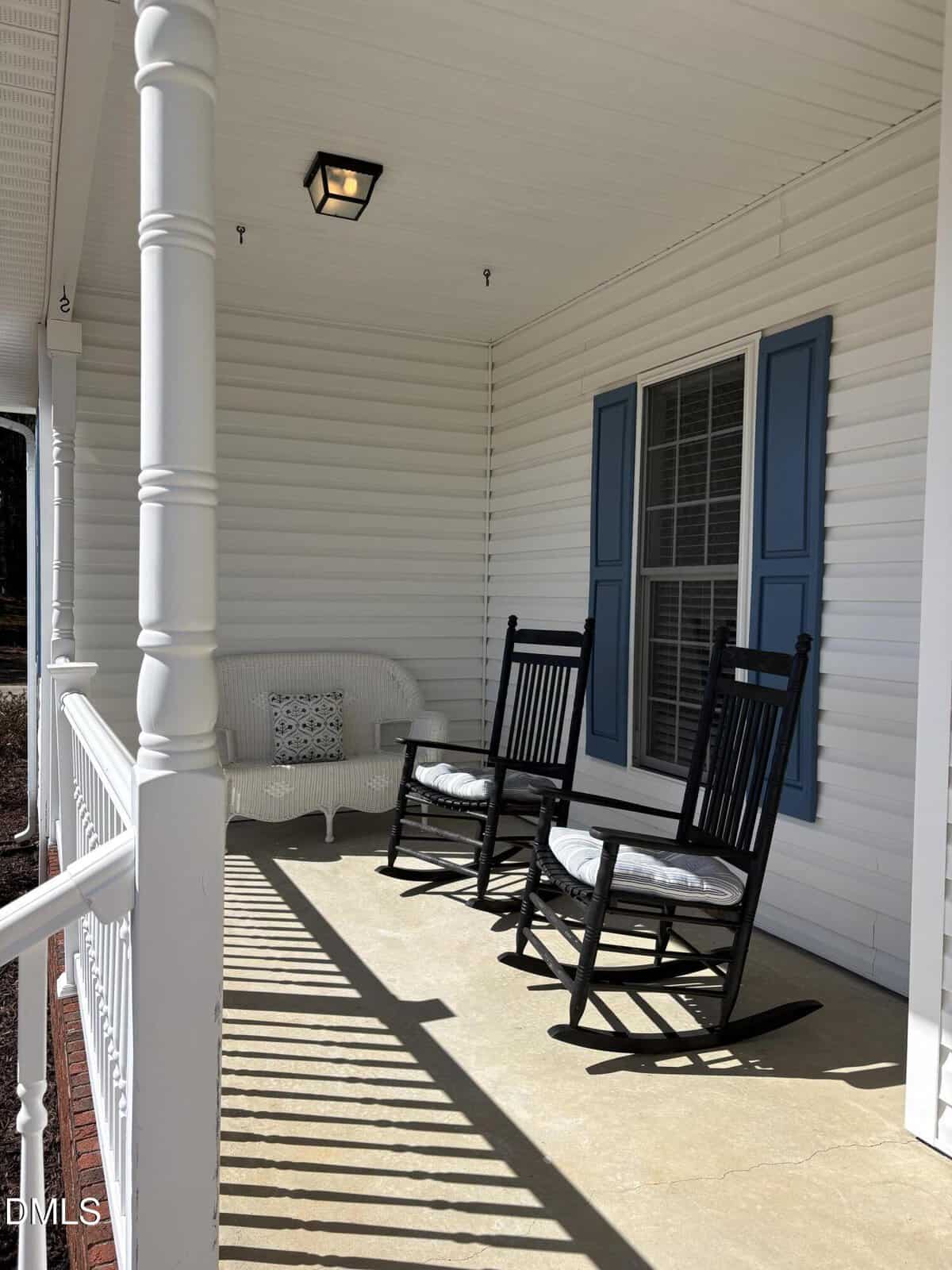 A sunny front porch with white railing, two black rocking chairs with cushions, a white wicker bench with a pillow, and blue shutters framing a large window. Shadows from the railing fall across the cement floor.