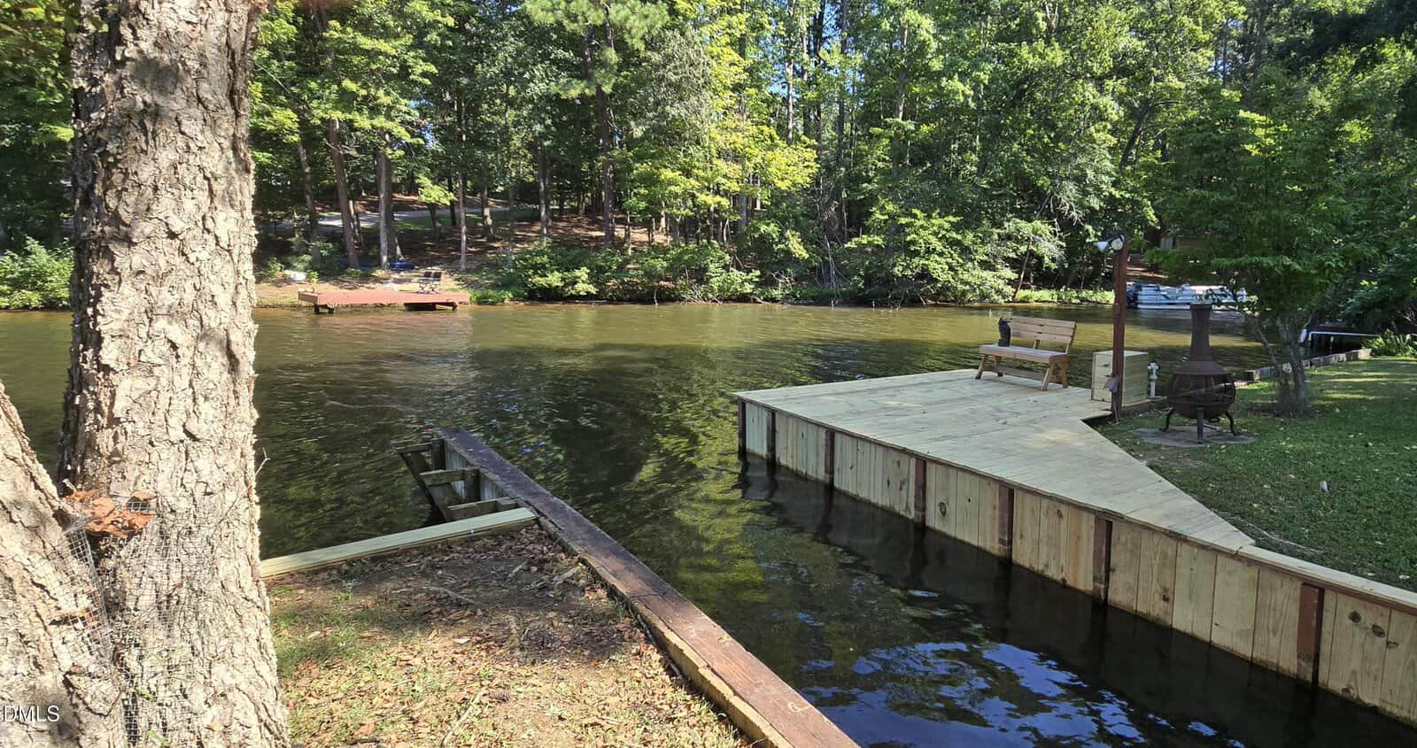 A wooden dock extends over a calm lake surrounded by trees, with a bench and fire pit nearby. Another small dock is visible across the water. Sunlight filters through the greenery.