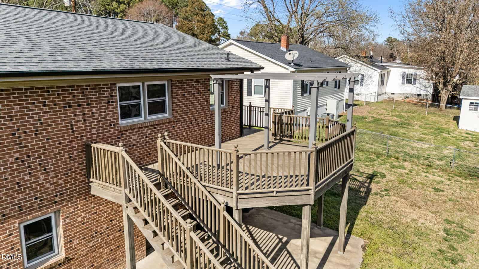 A wooden deck with stairs is attached to a brick house. The deck overlooks a grassy yard and is surrounded by other houses. The scene is set during daytime with clear skies.